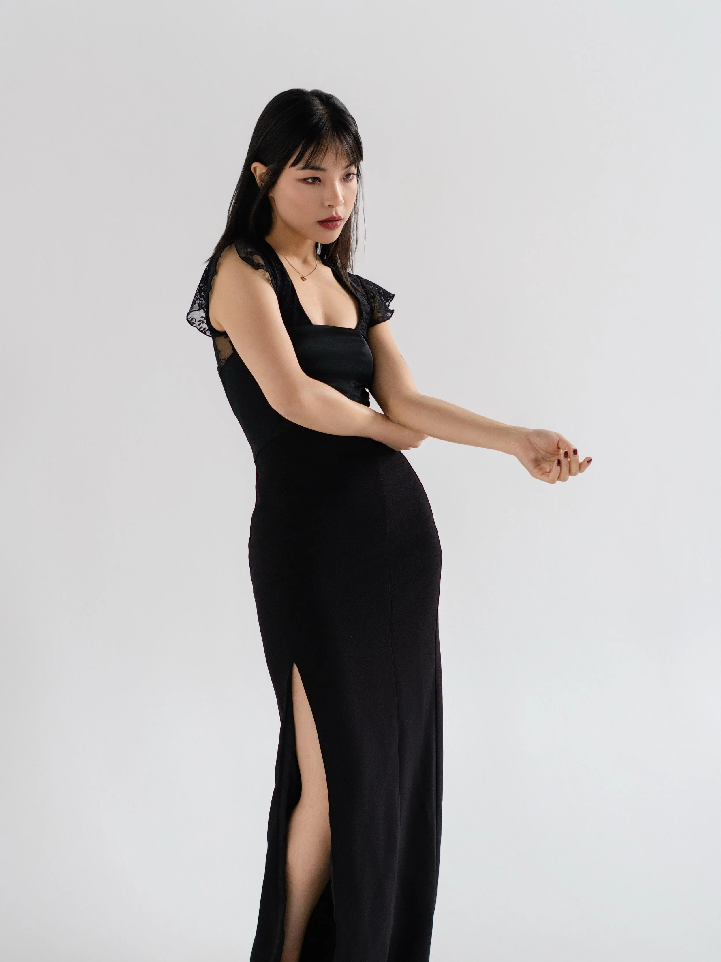 A woman in a black dress with lace details, standing against a white background, adjusting her sleeve.