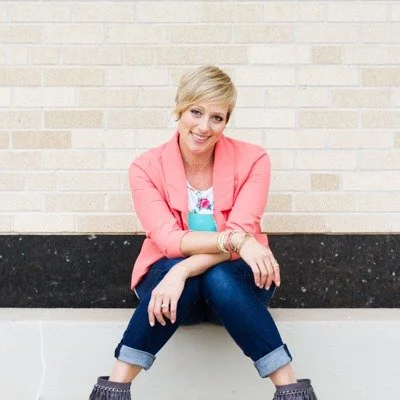 Person in a coral blazer and jeans sitting against a brick wall, smiling.