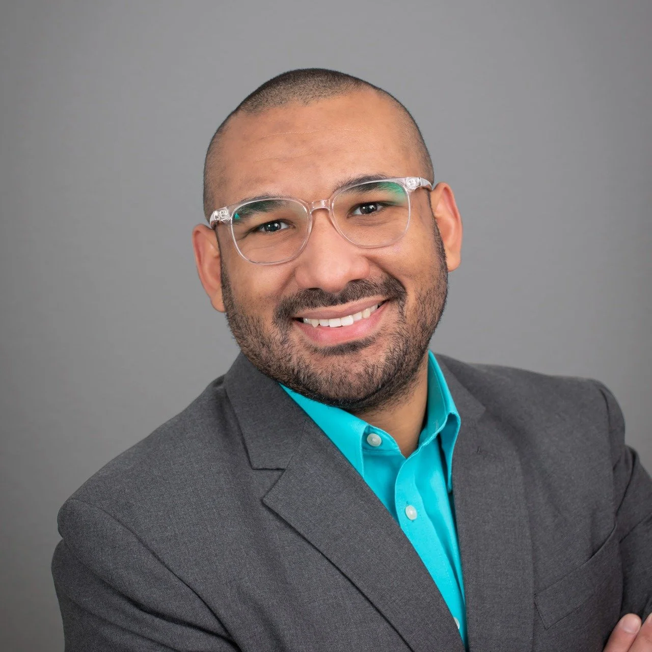 A smiling person wearing glasses and a gray suit with a teal shirt, against a gray background.