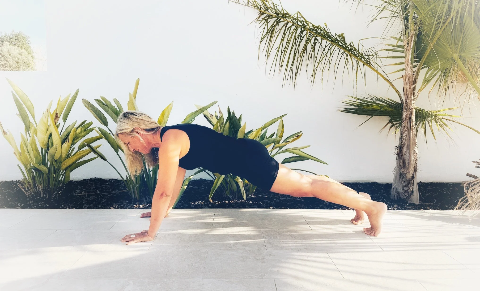 A woman practicing yoga outdoors in a low lunge position, surrounded by tropical plants and palm trees, with a white wall in the background.