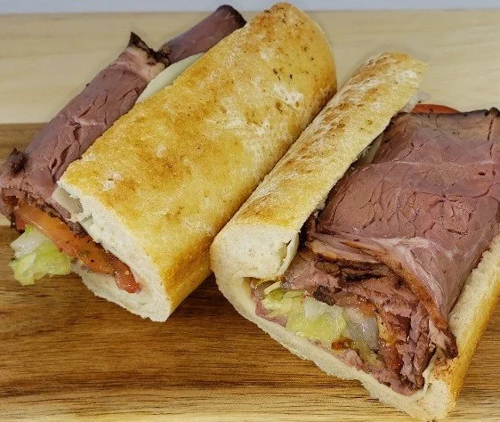 Two roast beef sandwiches with lettuce and tomato on hoagie buns, sliced in half and placed on a wooden surface.