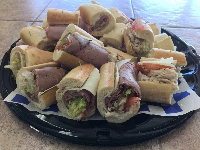 Assorted mini sliced sandwiches with meats, lettuce, and cheese on a black platter lined with paper, placed on a tiled surface.