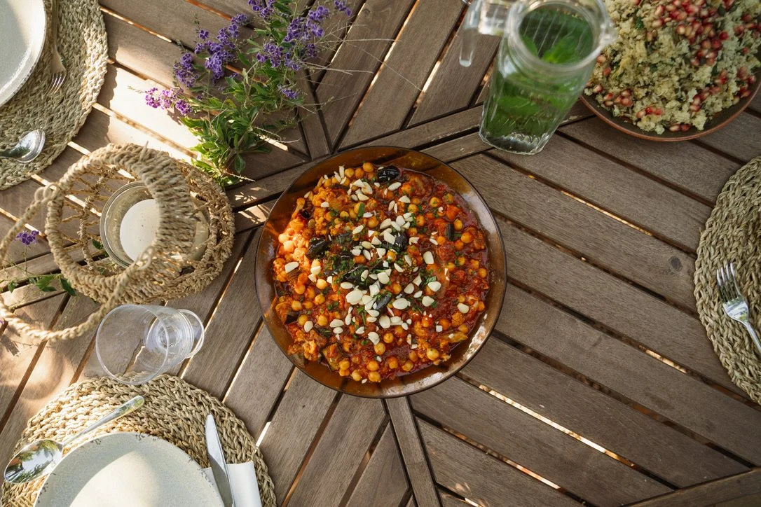 A wooden outdoor table set with woven placemats, white plates, silverware, a bowl of chickpea stew topped with sliced almonds, a glass of water with mint, a small lantern, a bouquet of purple flowers, and a dish of salad with pomegranate seeds.
