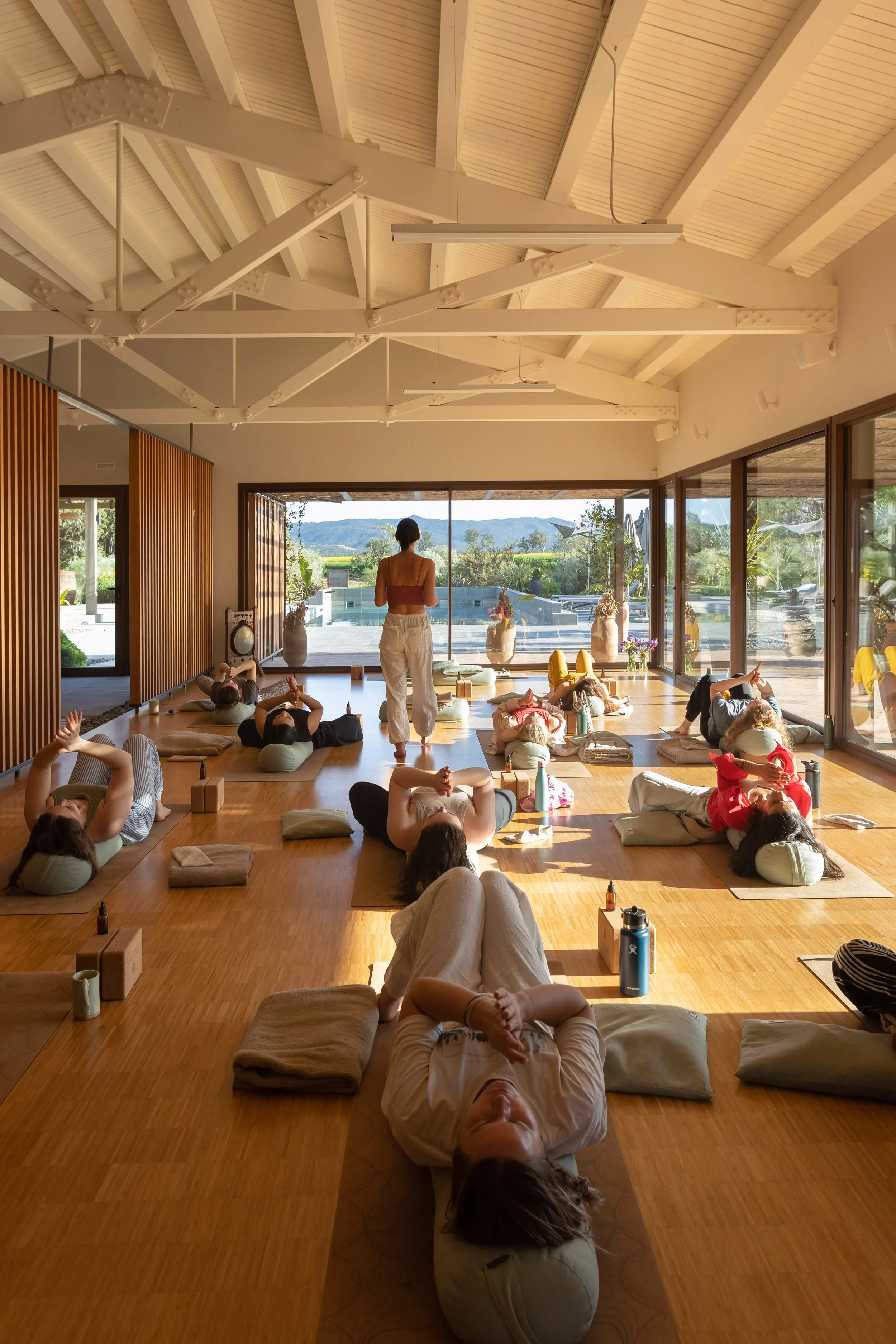 People participating in a yoga or meditation class in a bright, spacious room with large windows and wooden floors, some lying on mats and pillows, with an instructor at the front.