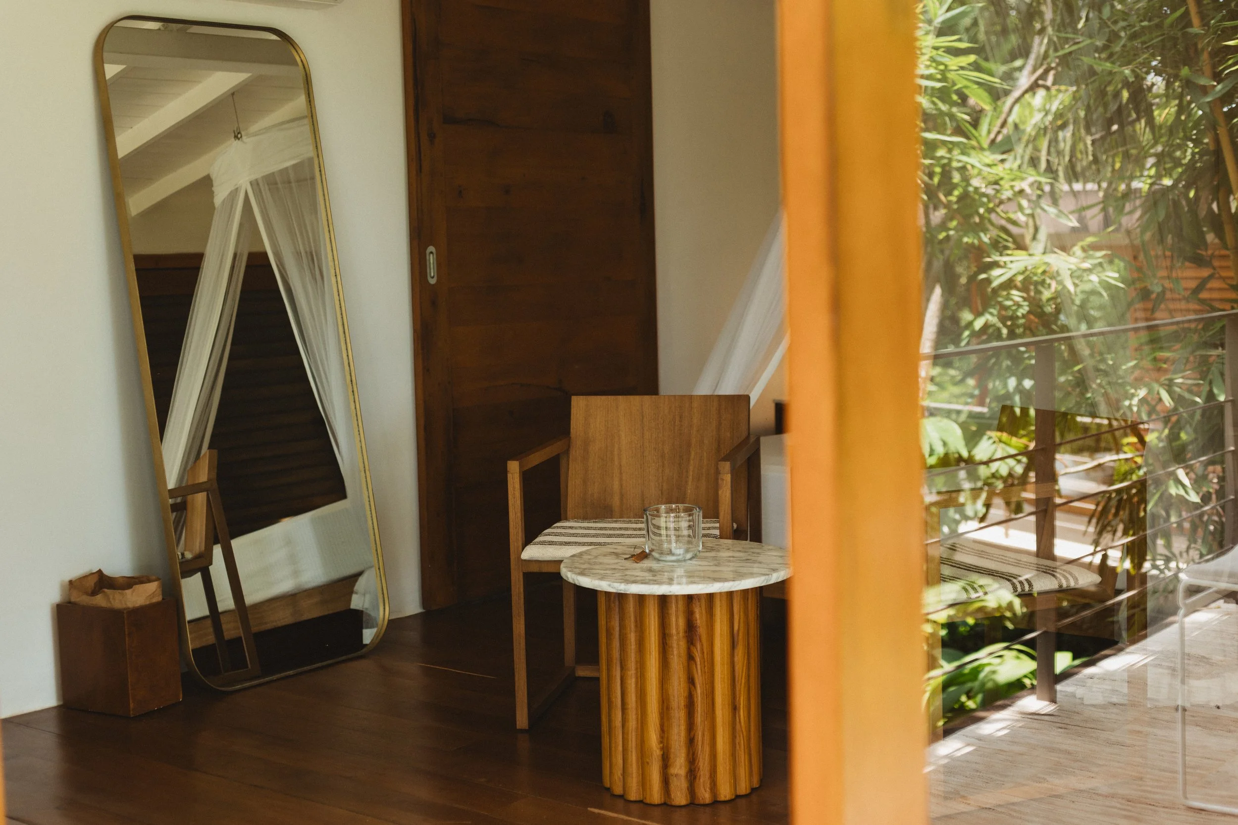 An interior view of a room with wooden furniture, a tall mirror leaning against the wall reflected in it, and a small round marble-topped table with a glass jar. Outside through a sliding glass door, green foliage is visible.