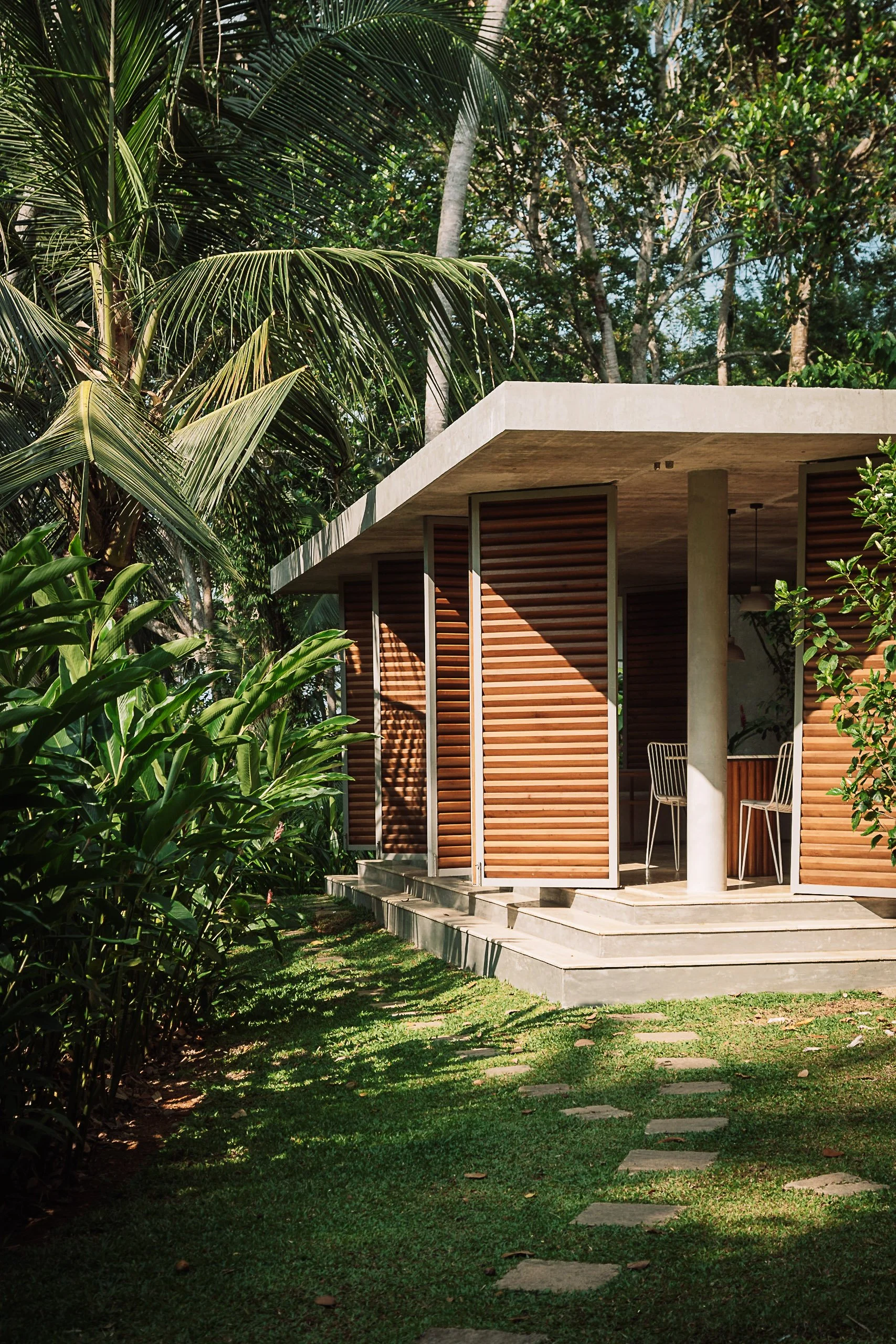 A modern house with a concrete roof and wooden slatted walls is situated among lush green tropical plants and tall trees, with a pathway leading to its steps.