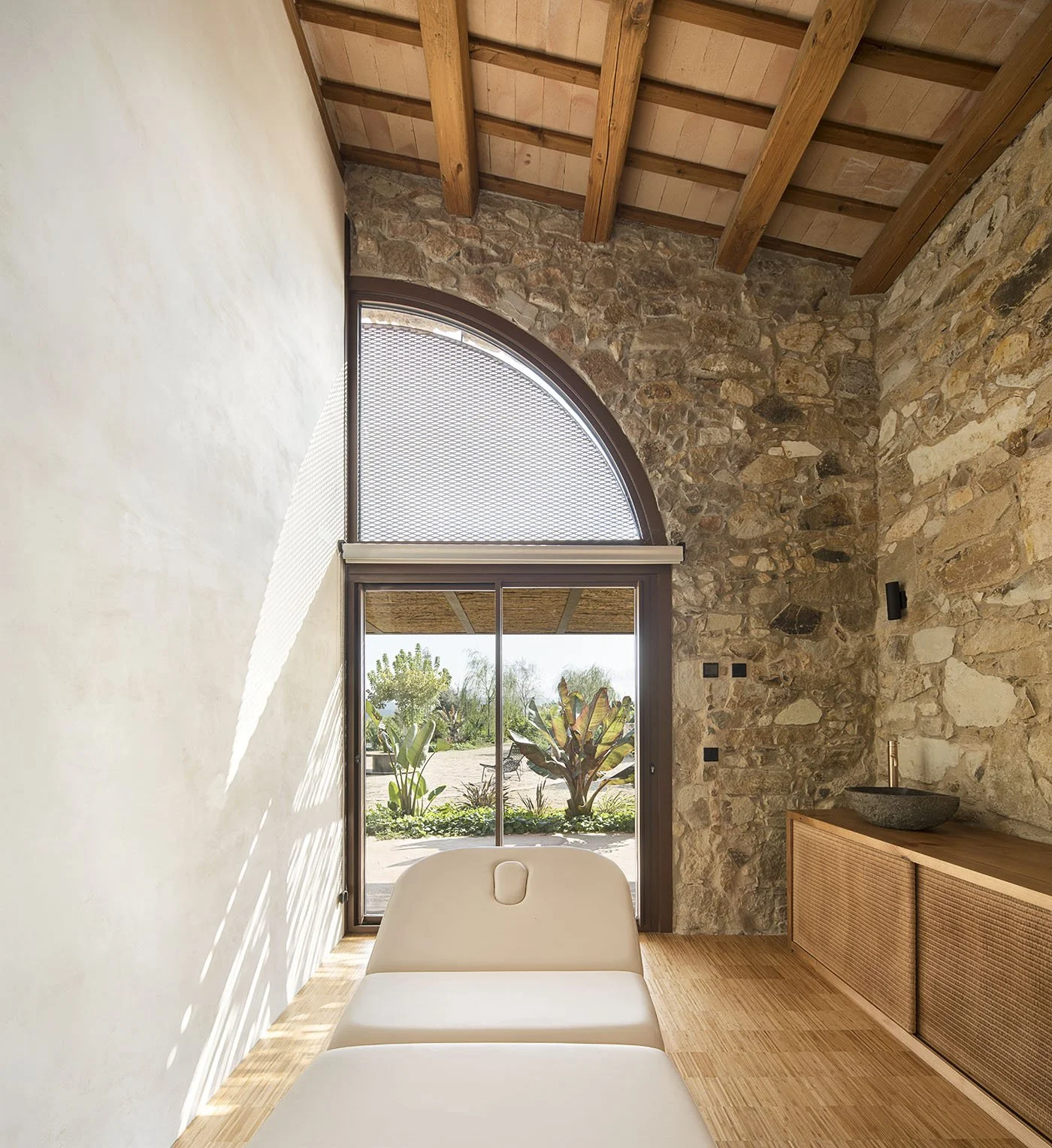 Interior of a room with stone walls, wooden ceiling with beams, a sliding glass door leading outside to greenery, and a cream-colored lounge chair.