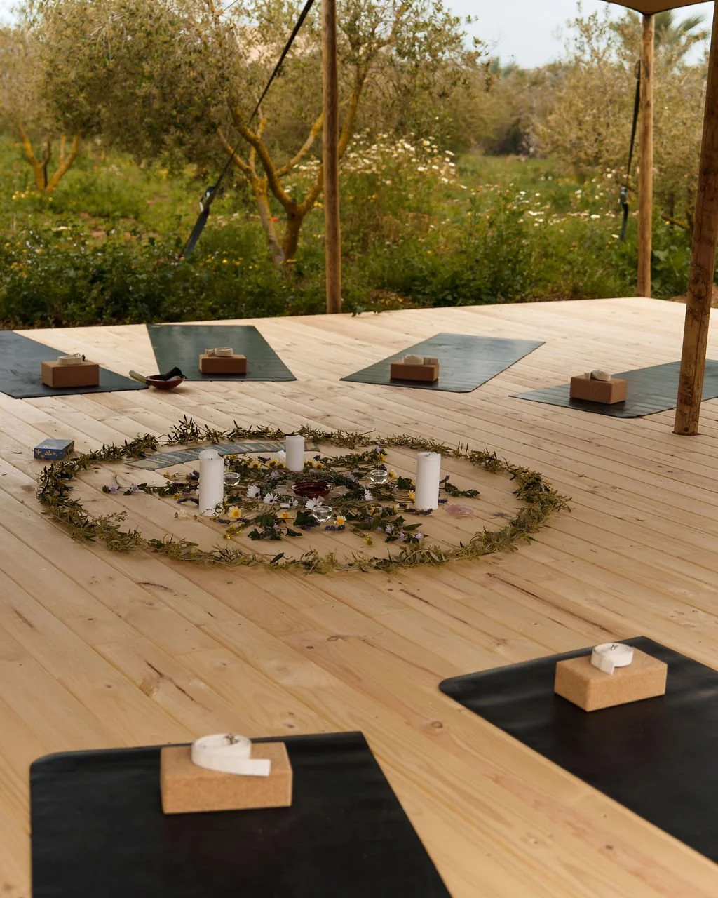 Indoor yoga or meditation space with black mats, cork blocks, and a decorative floral and candle centerpiece on a wooden floor, overlooking trees and a garden.
