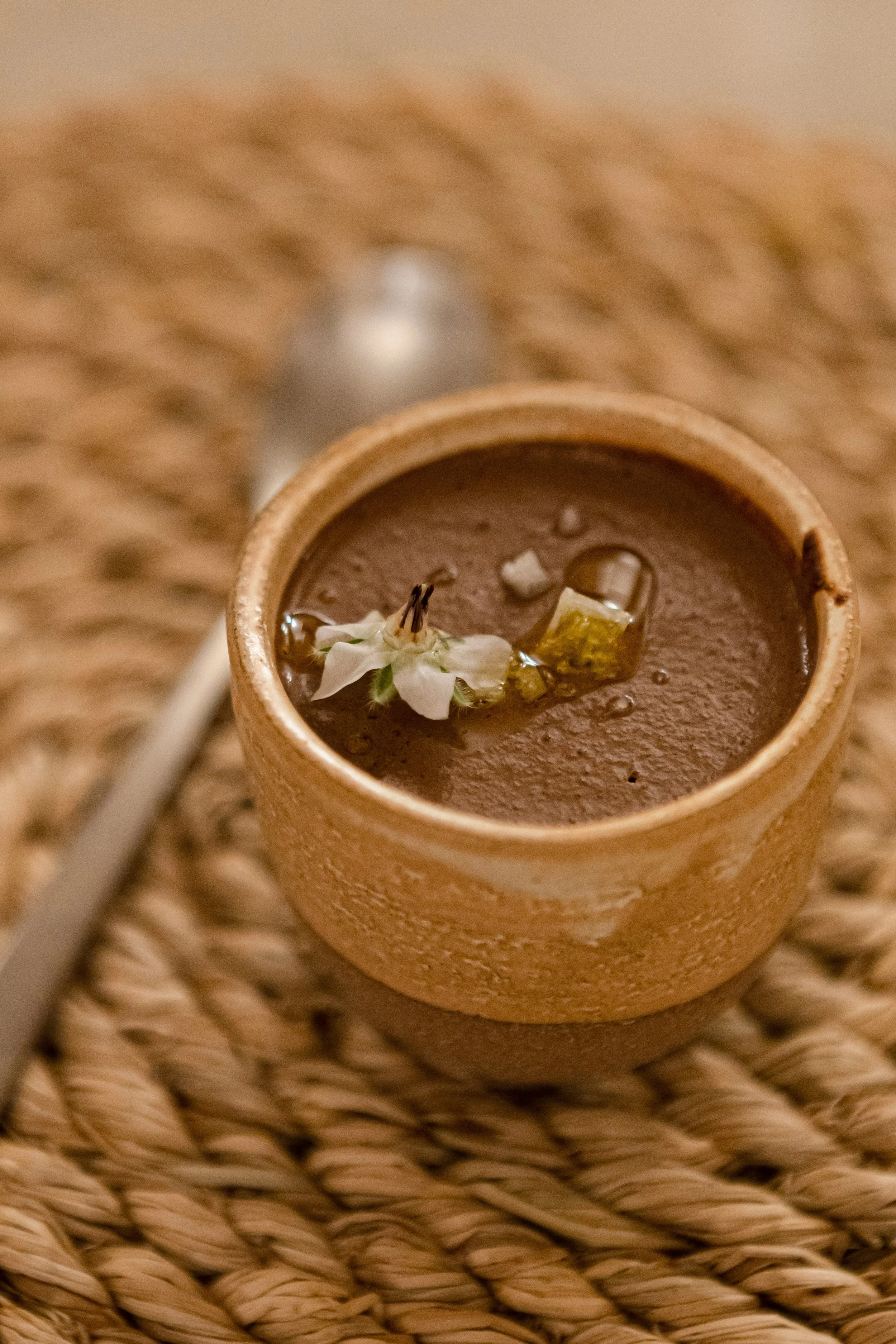A small, round, textured clay cup containing hot chocolate, garnished with a flower, a piece of lemon, and a small white item, resting on a woven rattan surface.