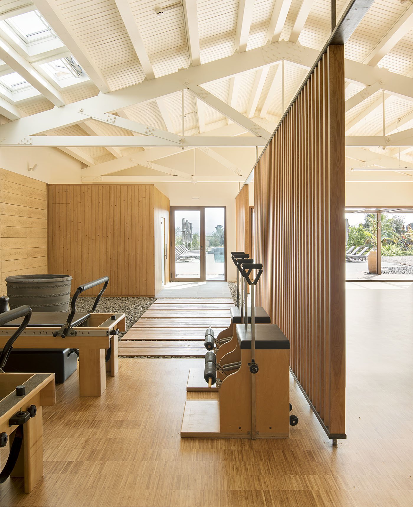 Indoor gym with wooden flooring, exercise machines, and a glass door leading outside to a sunny patio with lounge chairs.