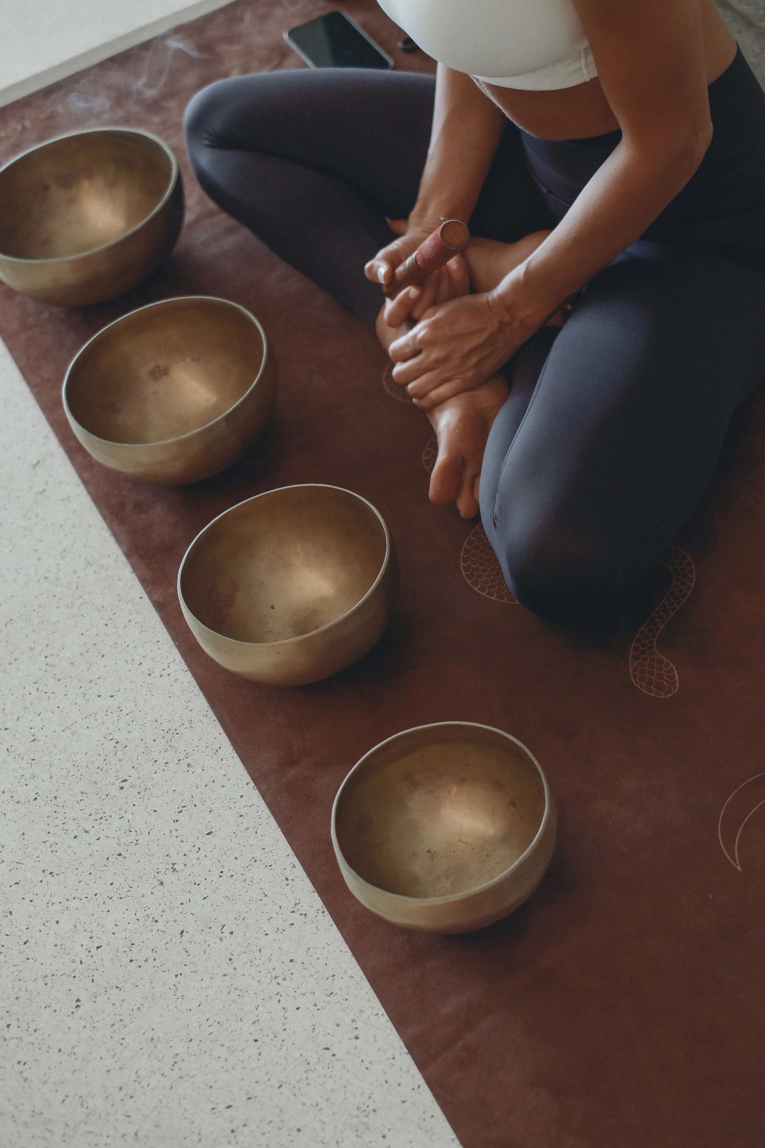 Person sitting cross-legged on the floor, playing singing bowls with a mallet. Four metallic singing bowls are arranged on a cloth in front of them.