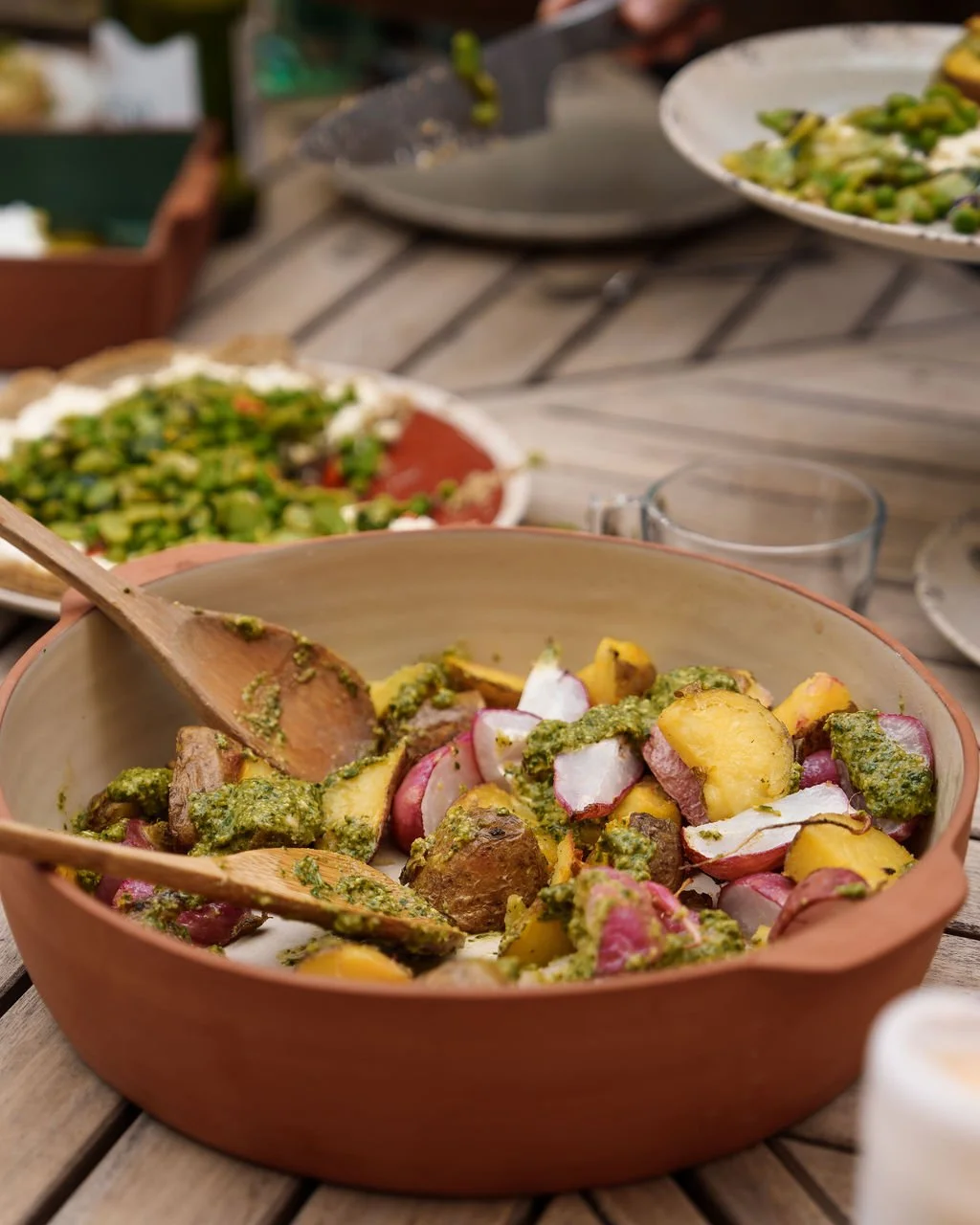 Salad with radishes, potatoes, and green sauce in a bowl on wooden table with other dishes in the background.
