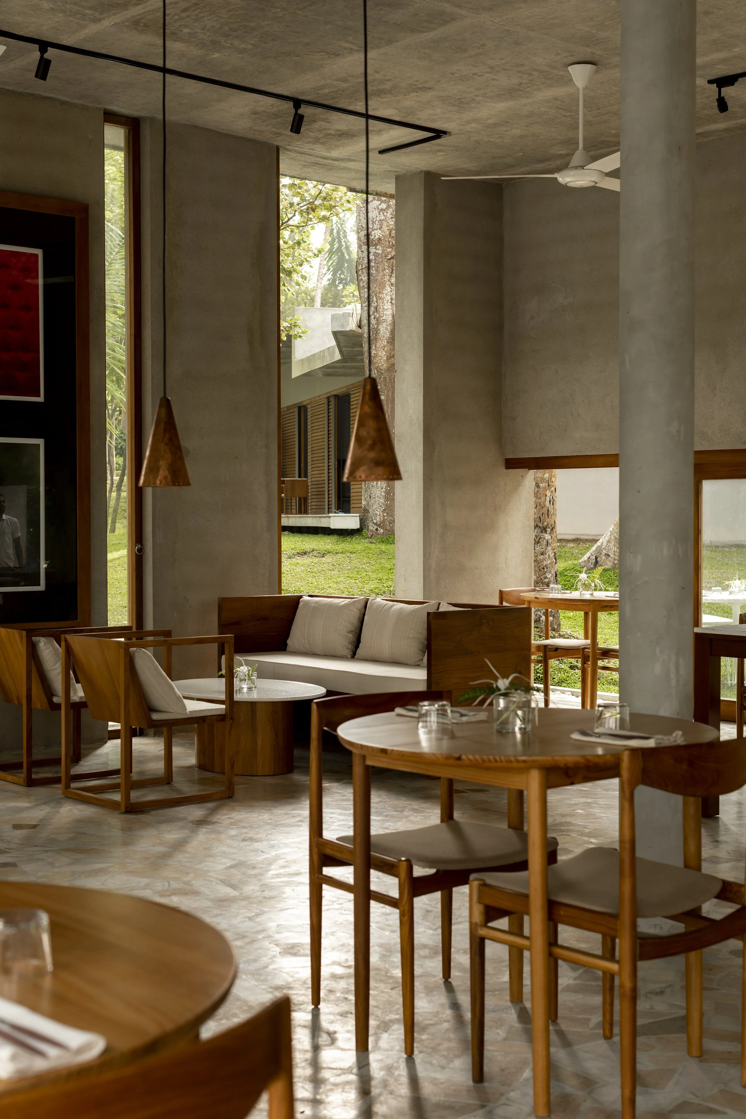 Interior of a modern restaurant with wooden tables, chairs, and a beige cushioned sofa, large windows showing greenery outside, and pendant lights hanging from a concrete ceiling.