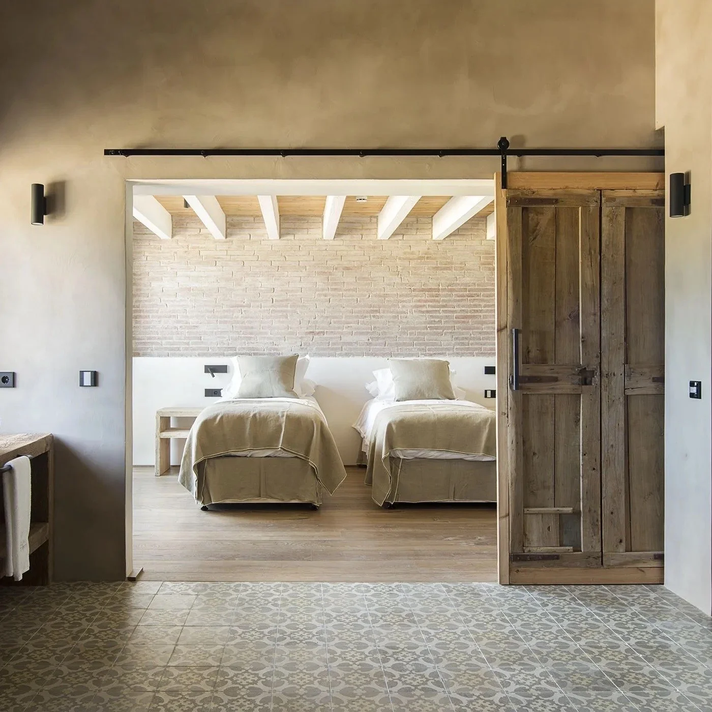 A bedroom with two single beds, beige bedding, and a brick accent wall, viewed through a doorway with a sliding barn door.
