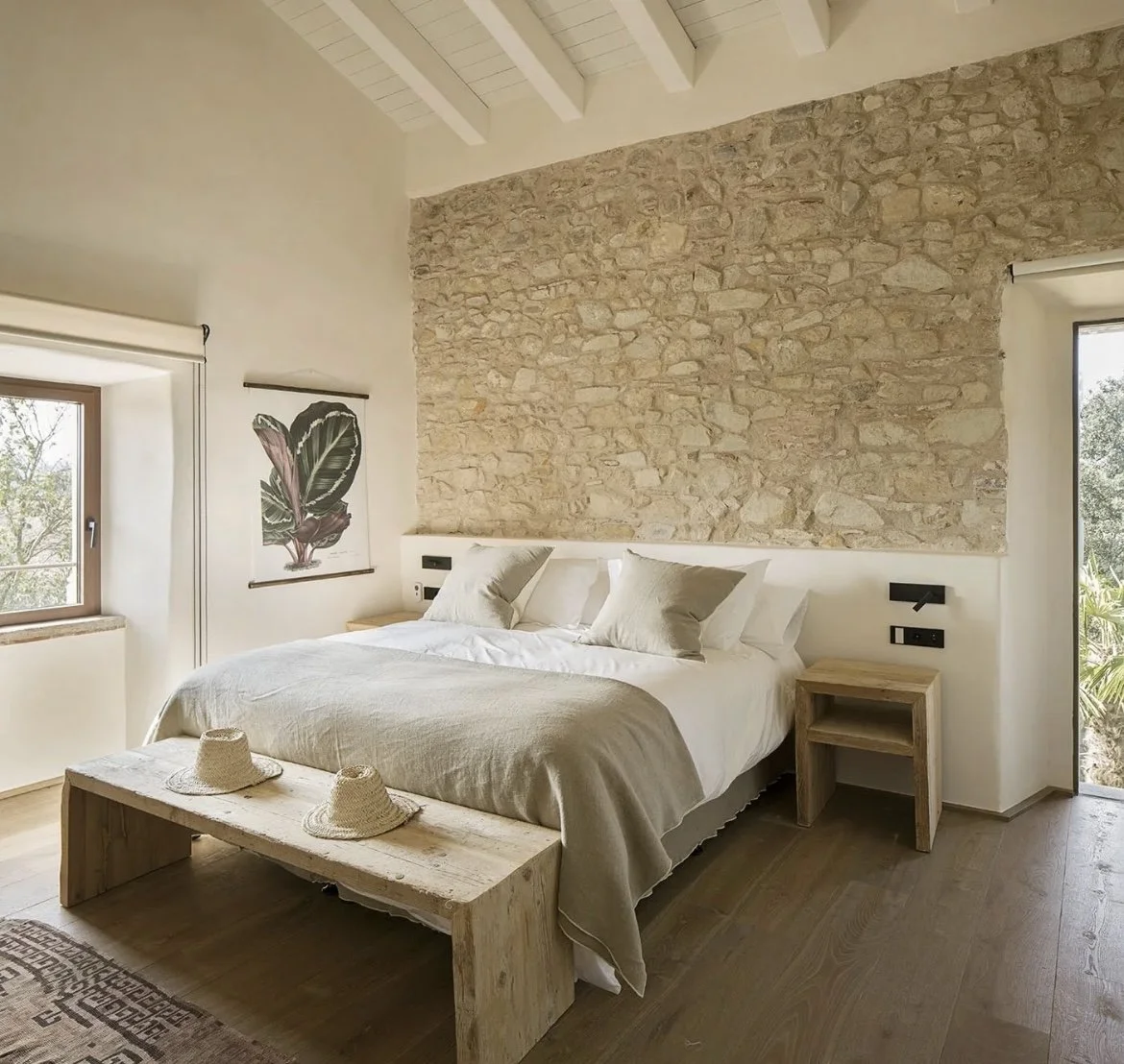 Bedroom with a stone accent wall, large bed with white bedding and pillows, wooden furniture including a bench at the foot of the bed and nightstands, a window with a view of trees, and a artwork of a leaf on the wall.