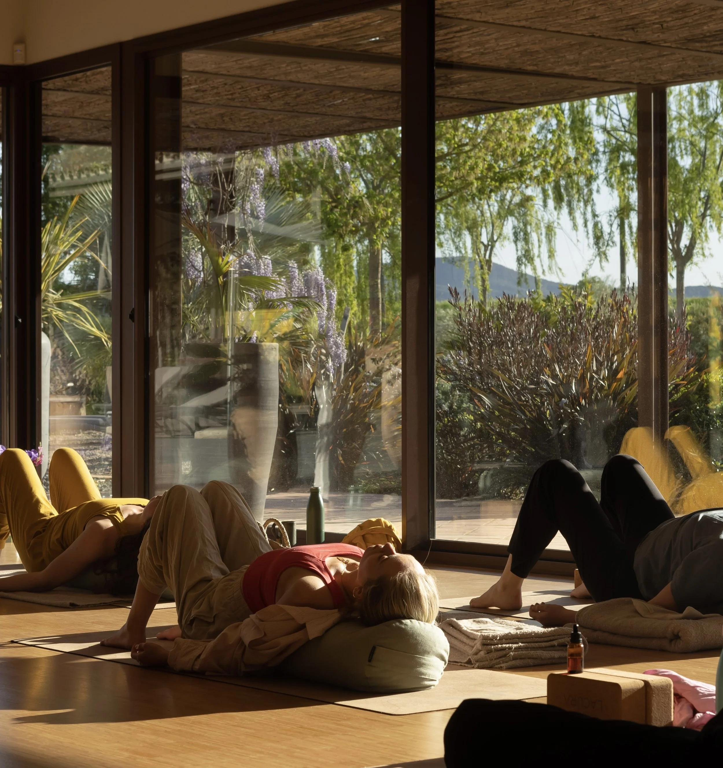 Group of people practicing yoga indoors, lying on mats with cushions, near large windows showing a garden view.