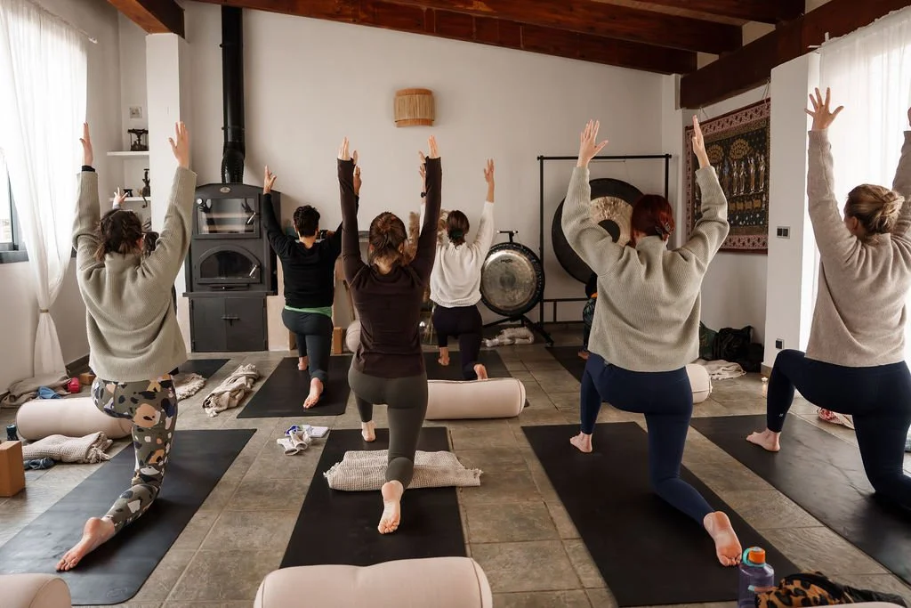 Group of people practicing yoga in a cozy, well-lit room with wooden beams, white curtains, and meditation accessories.