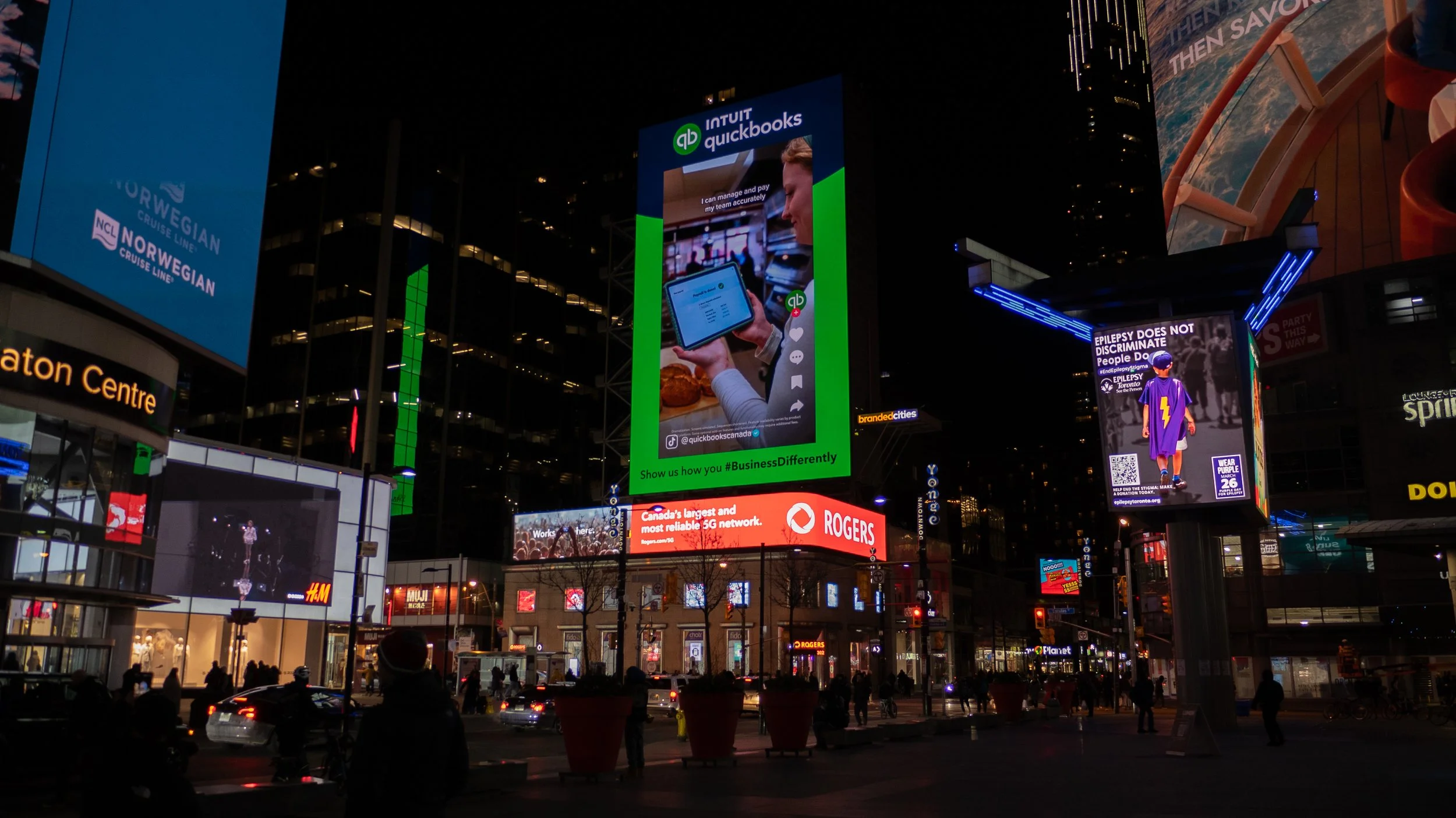 Night view of digital billboards in a bustling city center, featuring ads for Intuit QuickBooks, Norwegian Cruise Line, Rogers, epilepsy awareness, and more, with illuminated storefronts and pedestrians.
