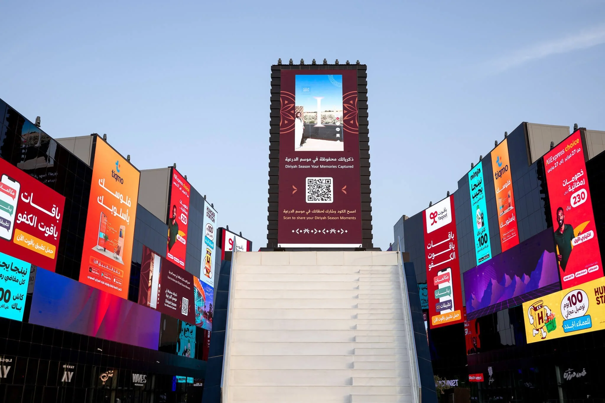 Outdoor digital advertising billboards in a commercial area with Arabic text, QR codes, and product promotions.