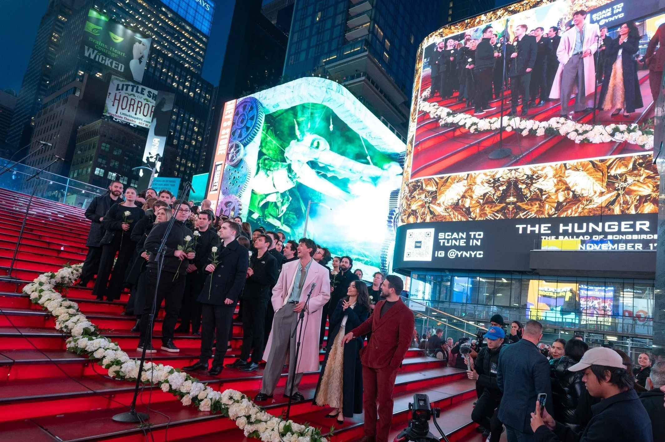 Group of people on red stairs in Times Square with large digital billboards and floral decorations at night.