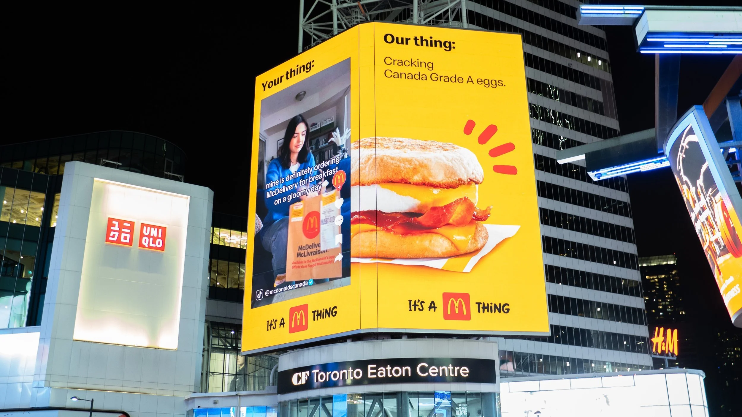 Large McDonald's breakfast sandwich billboard at night in Toronto Eaton Centre, next to Uniqlo logo.