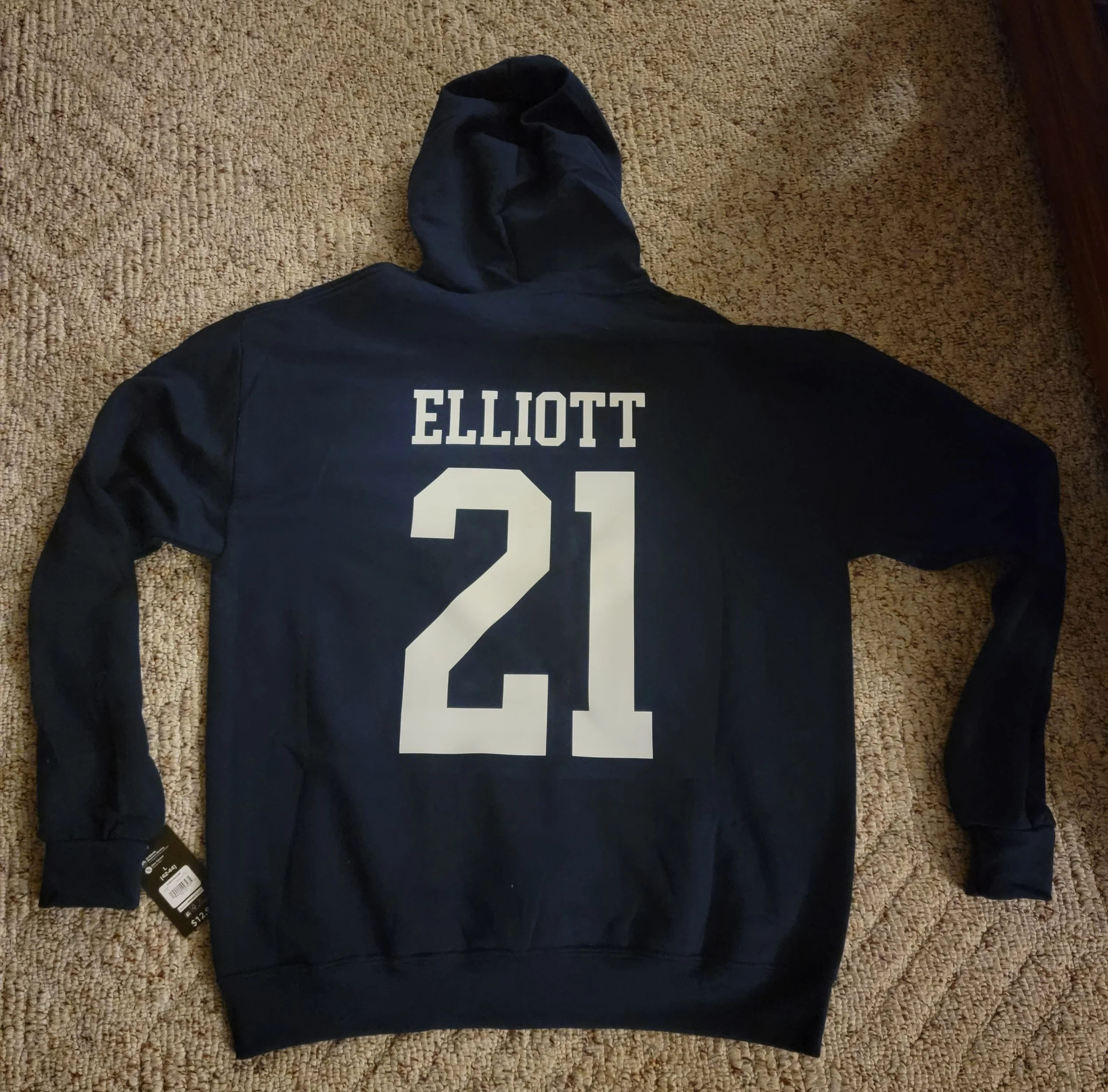 Black hoodie with the name 'ELLIOTT' and the number '21' printed on the back, laid flat on a beige carpet.