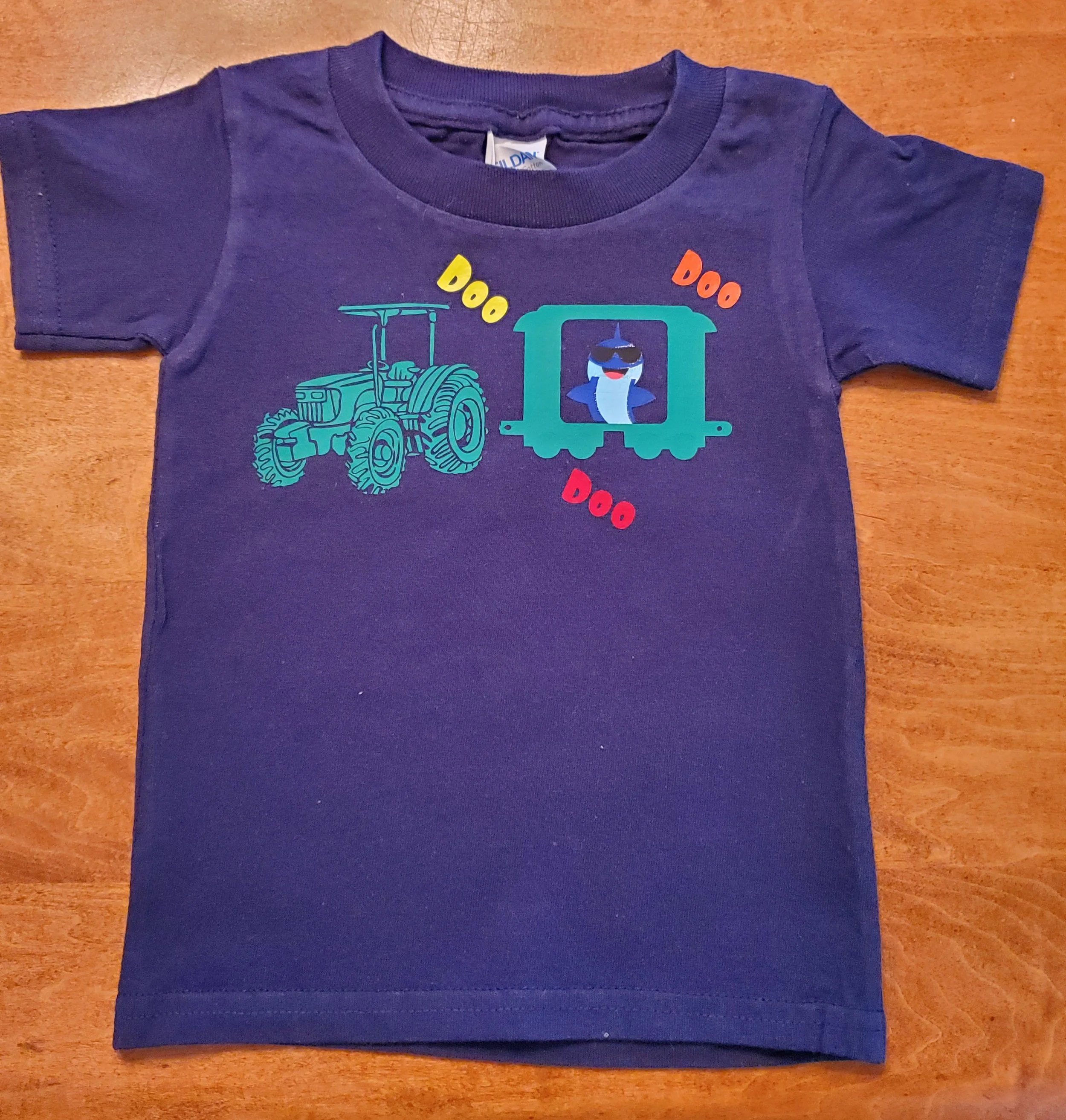 Blue children's t-shirt with a design of a tractor and a train car with a whale inside, and the words 'Doo Doo' in yellow, orange, and red.