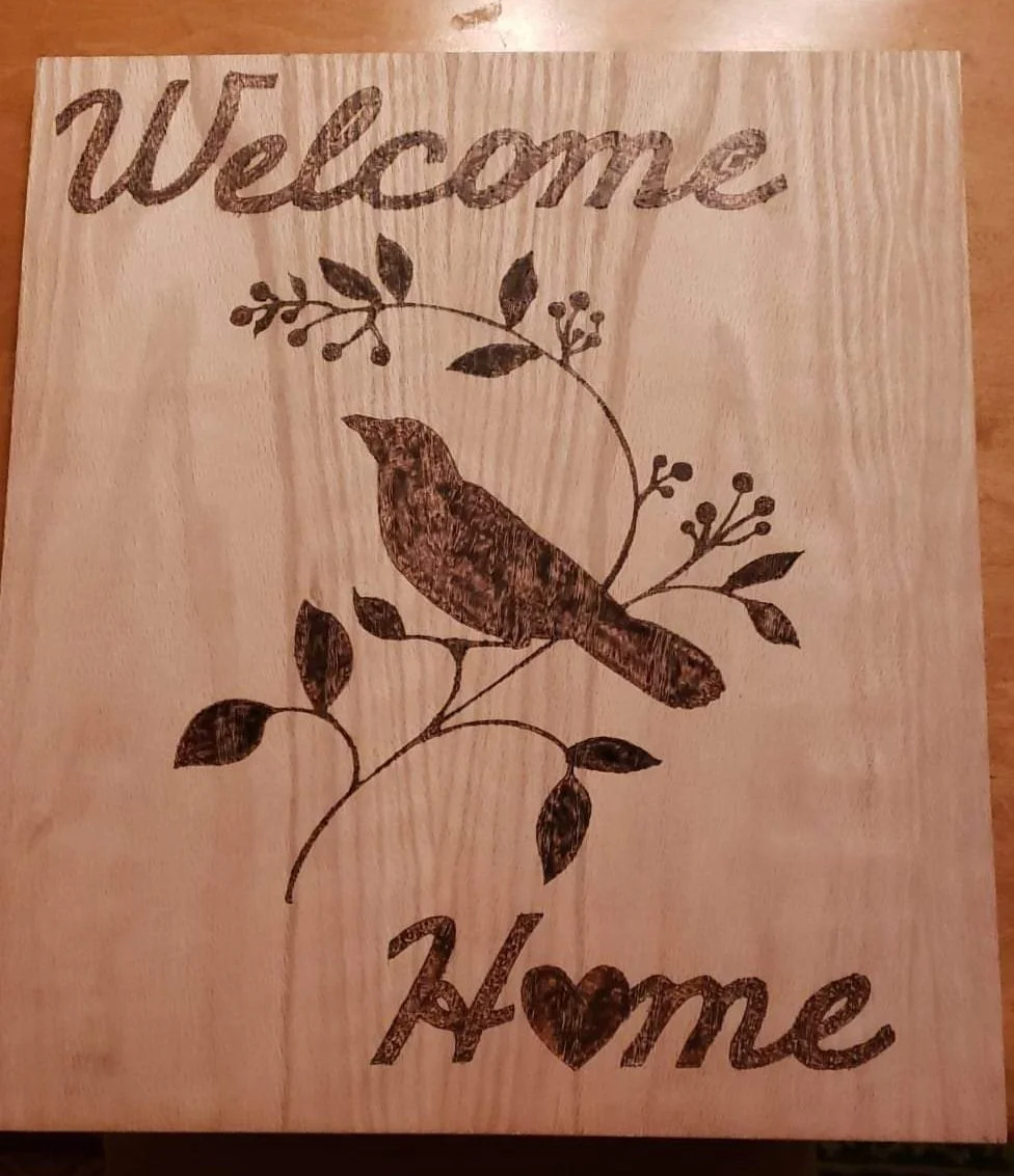 Wooden sign with the words 'Welcome' at the top and 'Home' at the bottom, featuring a bird sitting on a branch with leaves and berries.
