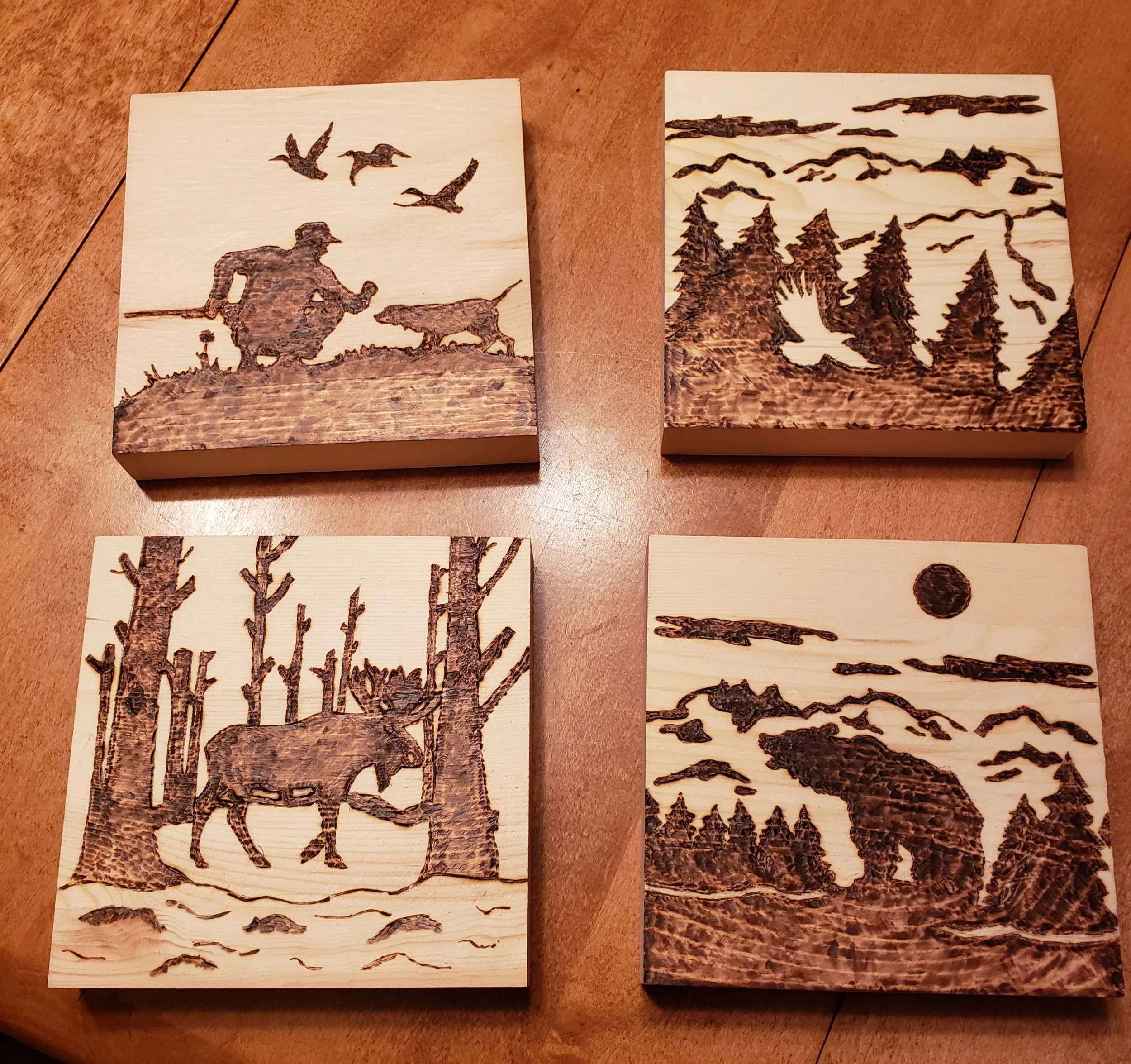 Set of four wooden plaques with burned images of outdoor wilderness scenes, including a hunter with a dog and birds, a mountain landscape with trees, a moose among trees, and a bear in the forest with a full moon.