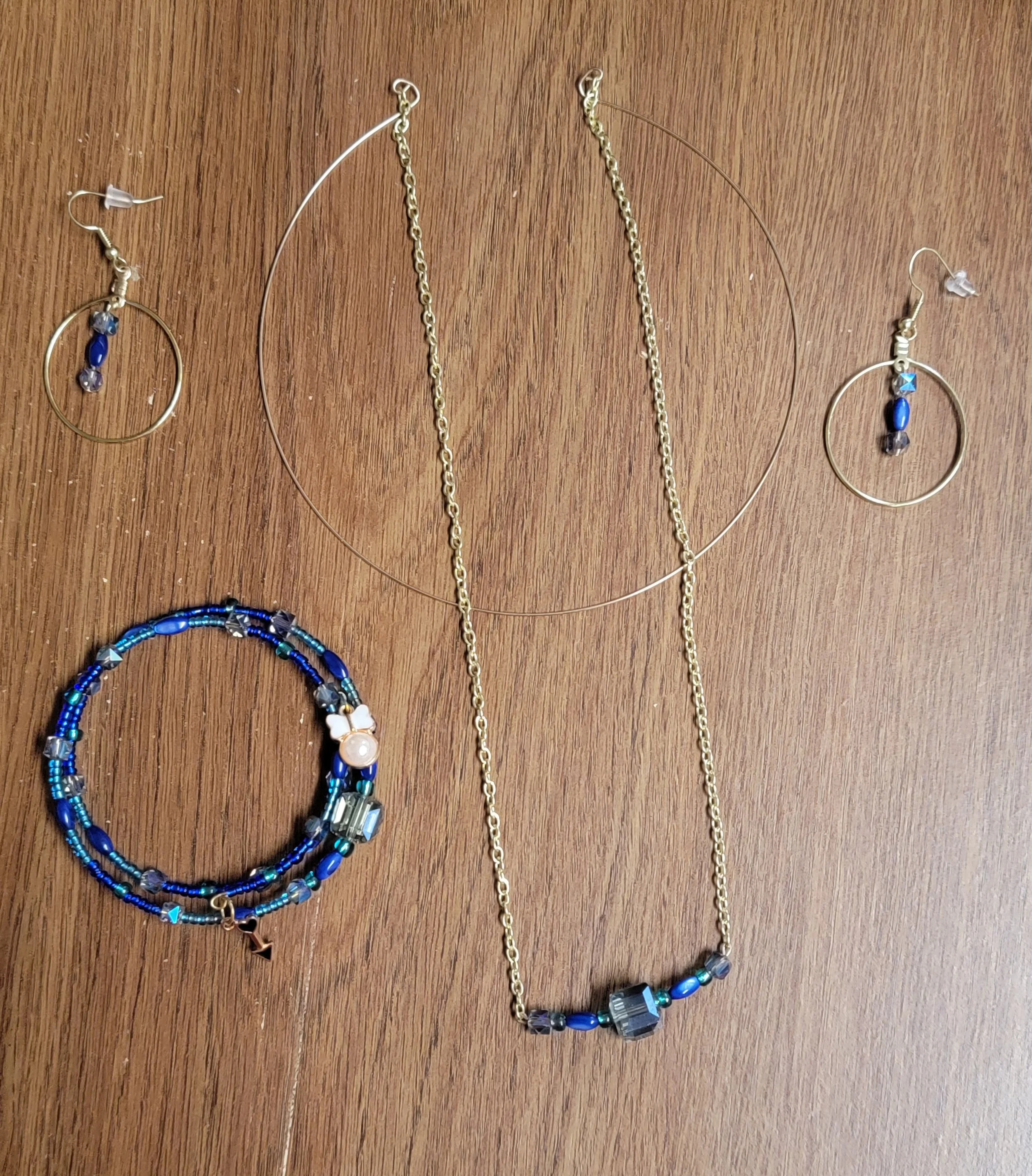 Set of jewelry including a pair of earrings, a necklace, and a bracelet, featuring dark blue and clear beads, with gold and silver accents, displayed on a wooden surface.