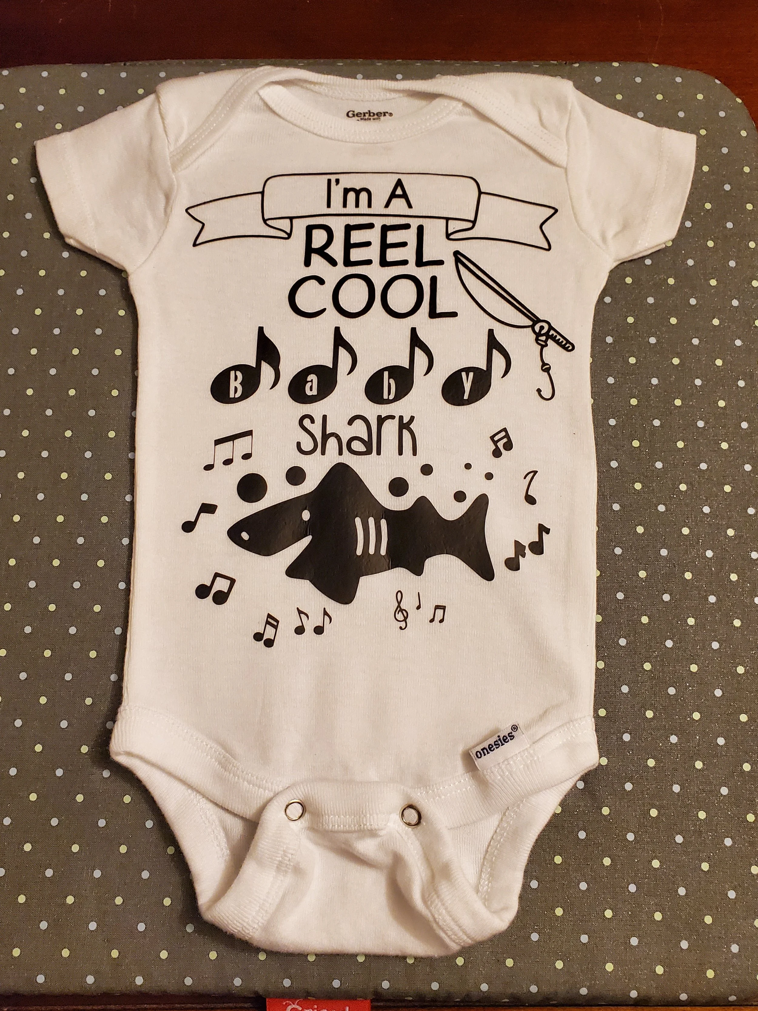 Baby onesie with the text "I'm a REEL COOL baby shark" and graphics of a fishing pole and shark surrounded by musical notes.