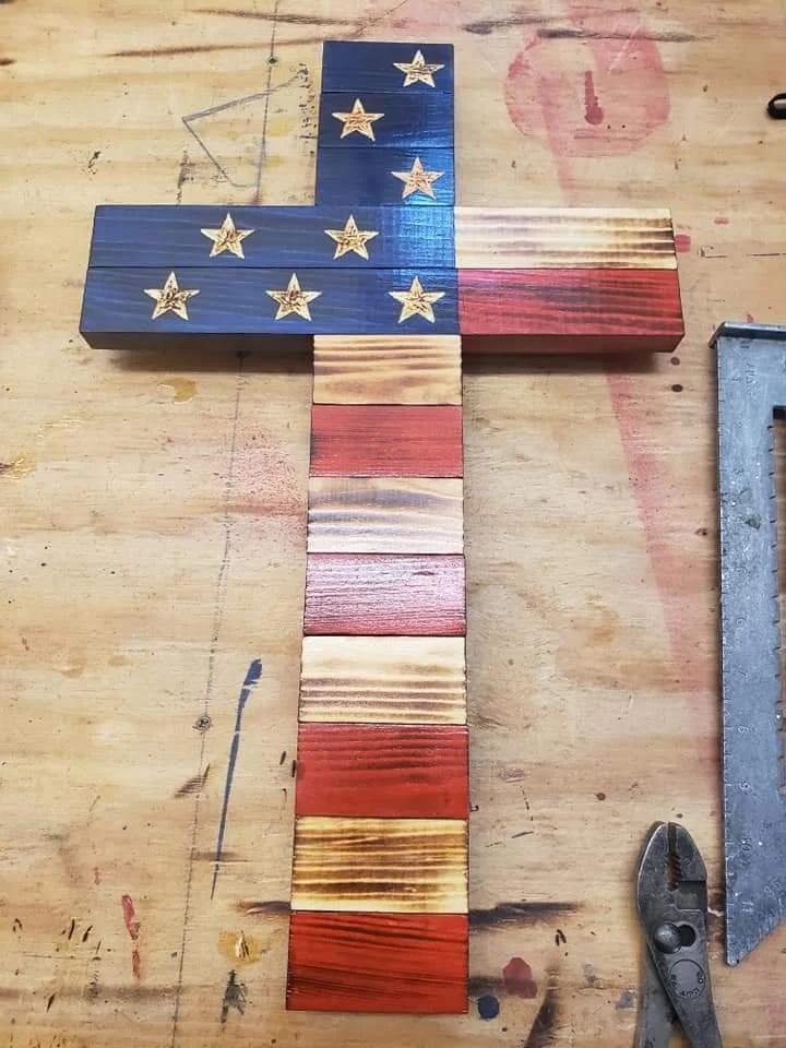 Wooden cross-shaped decoration painted with the American flag design, featuring blue with white stars at the top and red and beige stripes down the middle, placed on a work table with tools around.