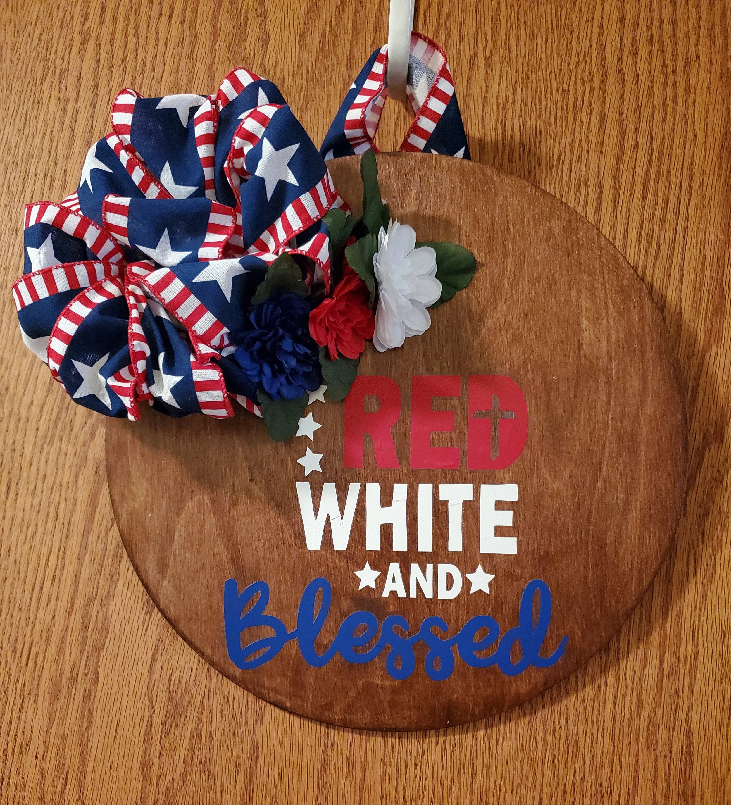 Decorative wooden plaque with red, white, and blue lettering that says "Red White and Blessed," adorned with an American flag-themed ribbon and white, red, and blue artificial flowers, hanging on a wooden surface.