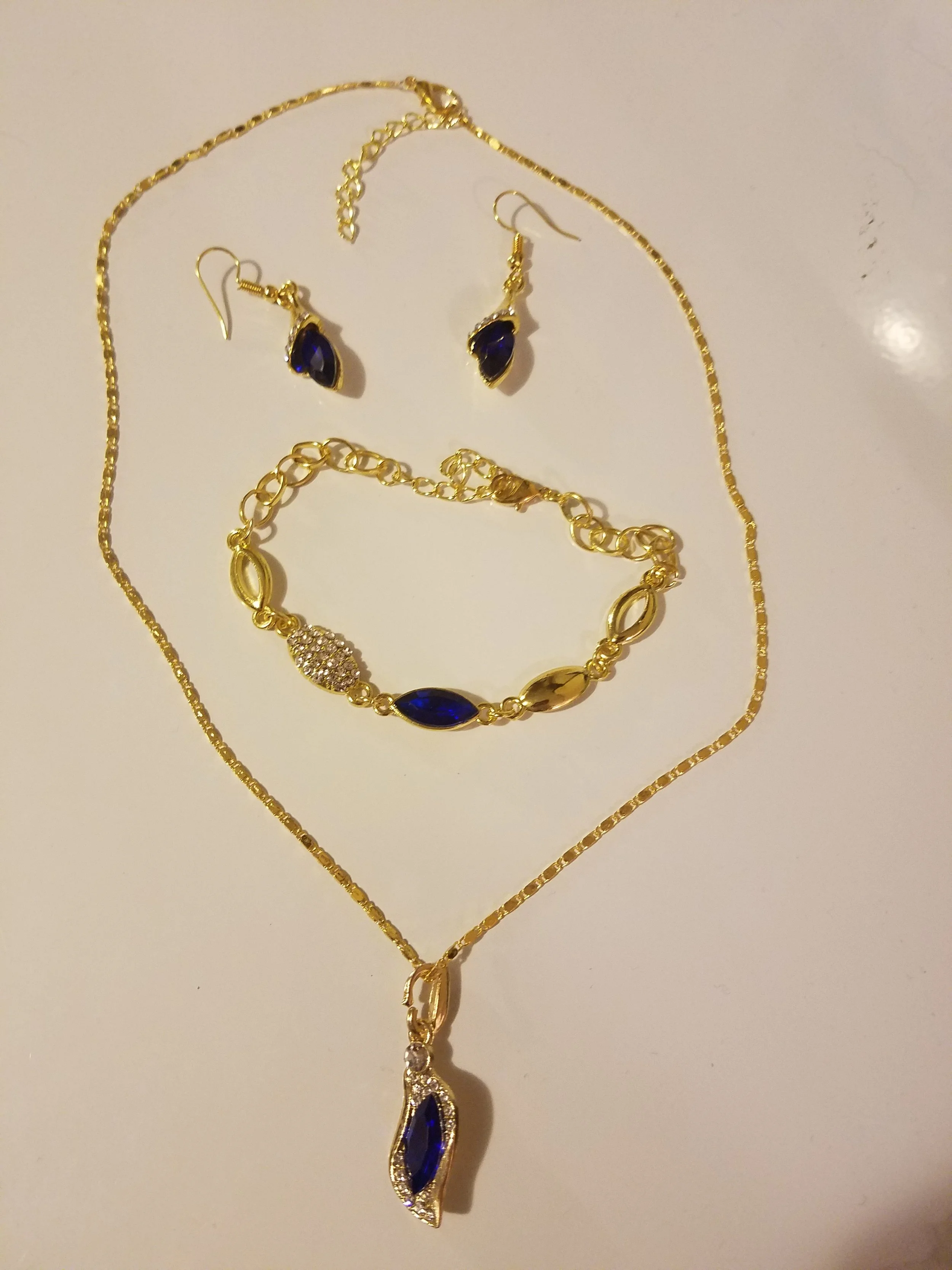 Gold necklace with blue gemstone pendant, matching gold and blue gemstone earrings, and a gold bracelet with blue and clear gemstones, laid out on a white surface.