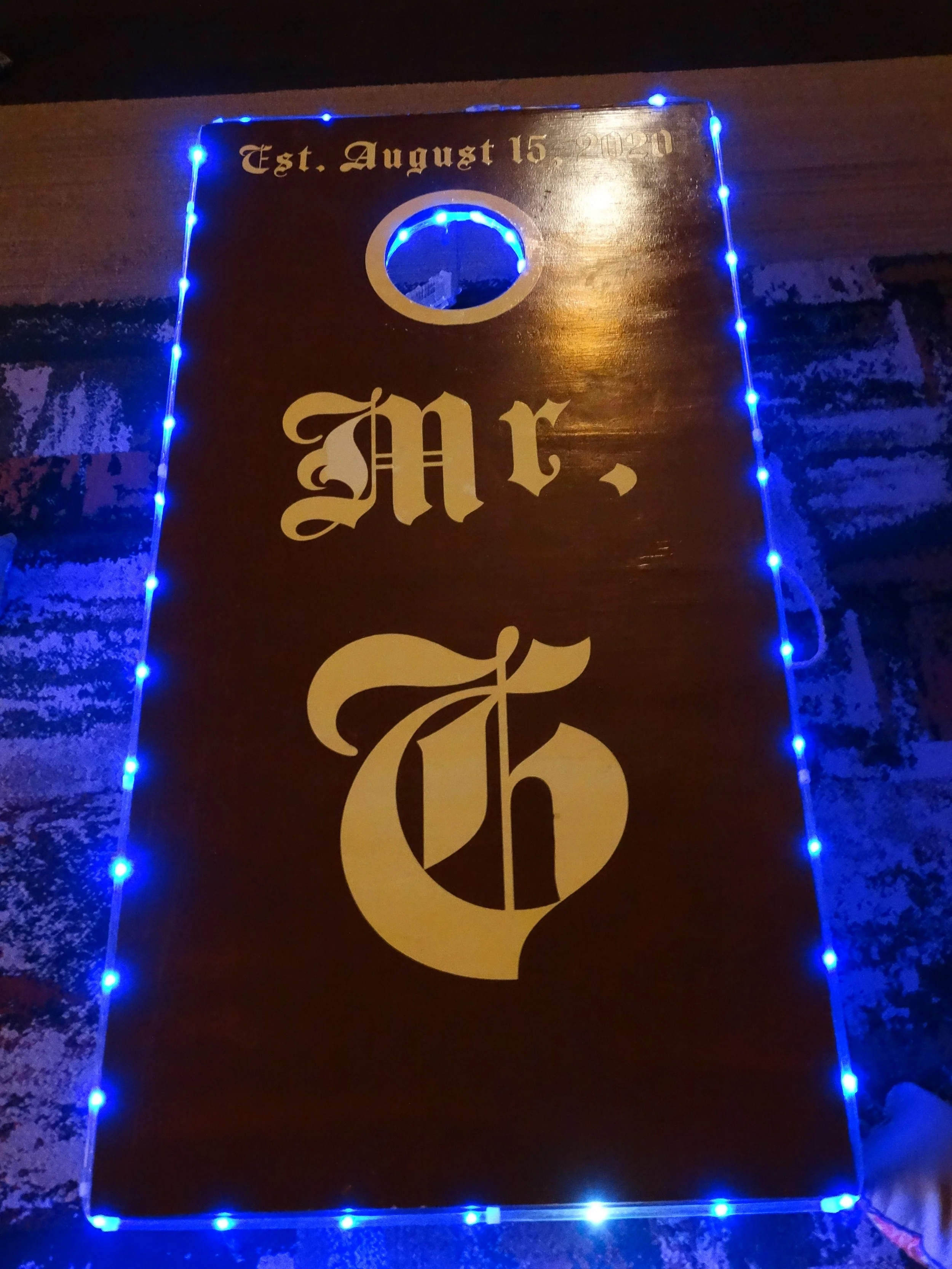 Wooden plaque with the word 'Mr.' in large decorative gold letters, surrounded by blue LED string lights. The top of the plaque reads 'Est. August 15 2020' in gold text.