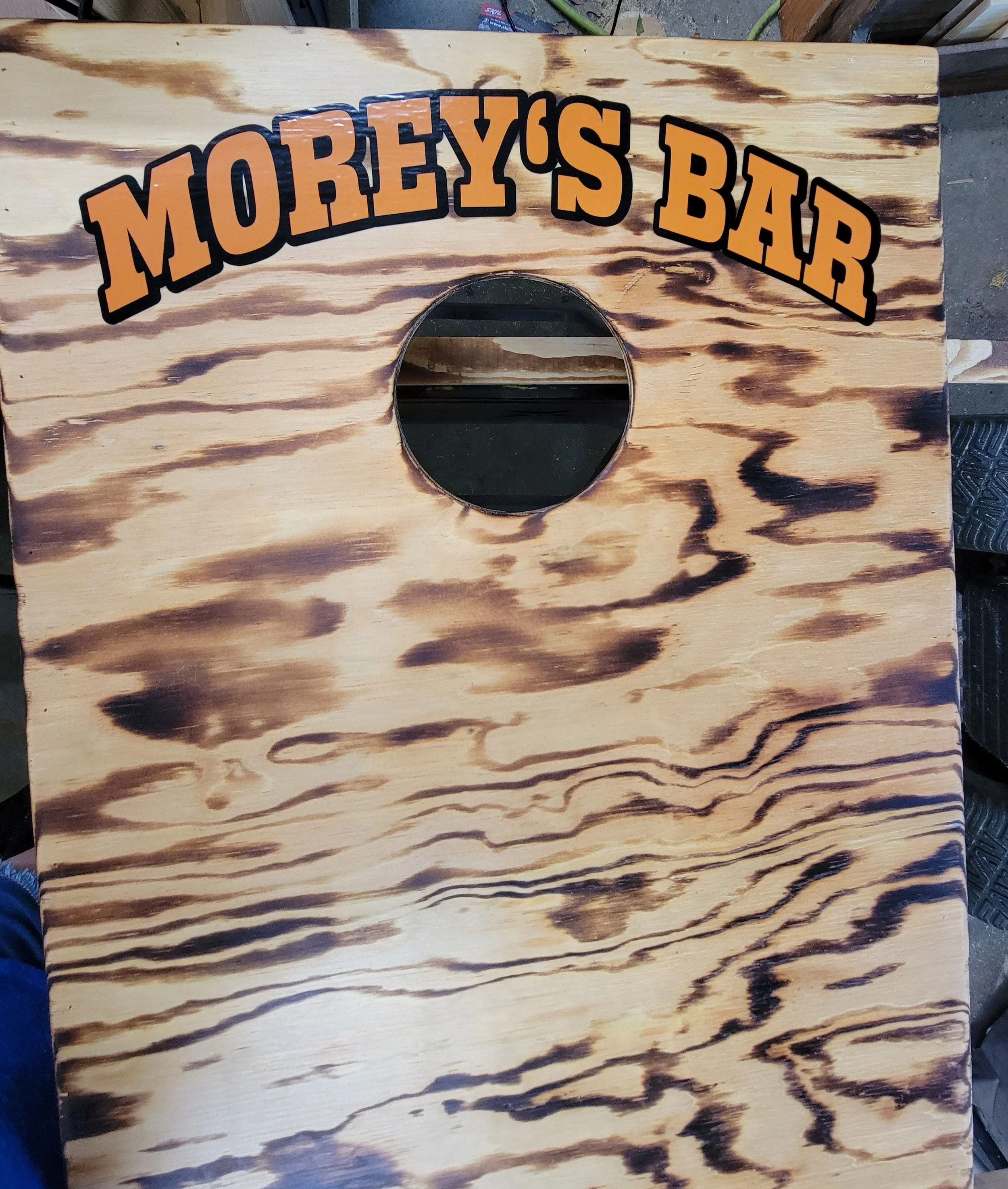 A wooden cornhole game board with the text 'Morey's Bar' painted at the top, featuring a circular hole near the center.