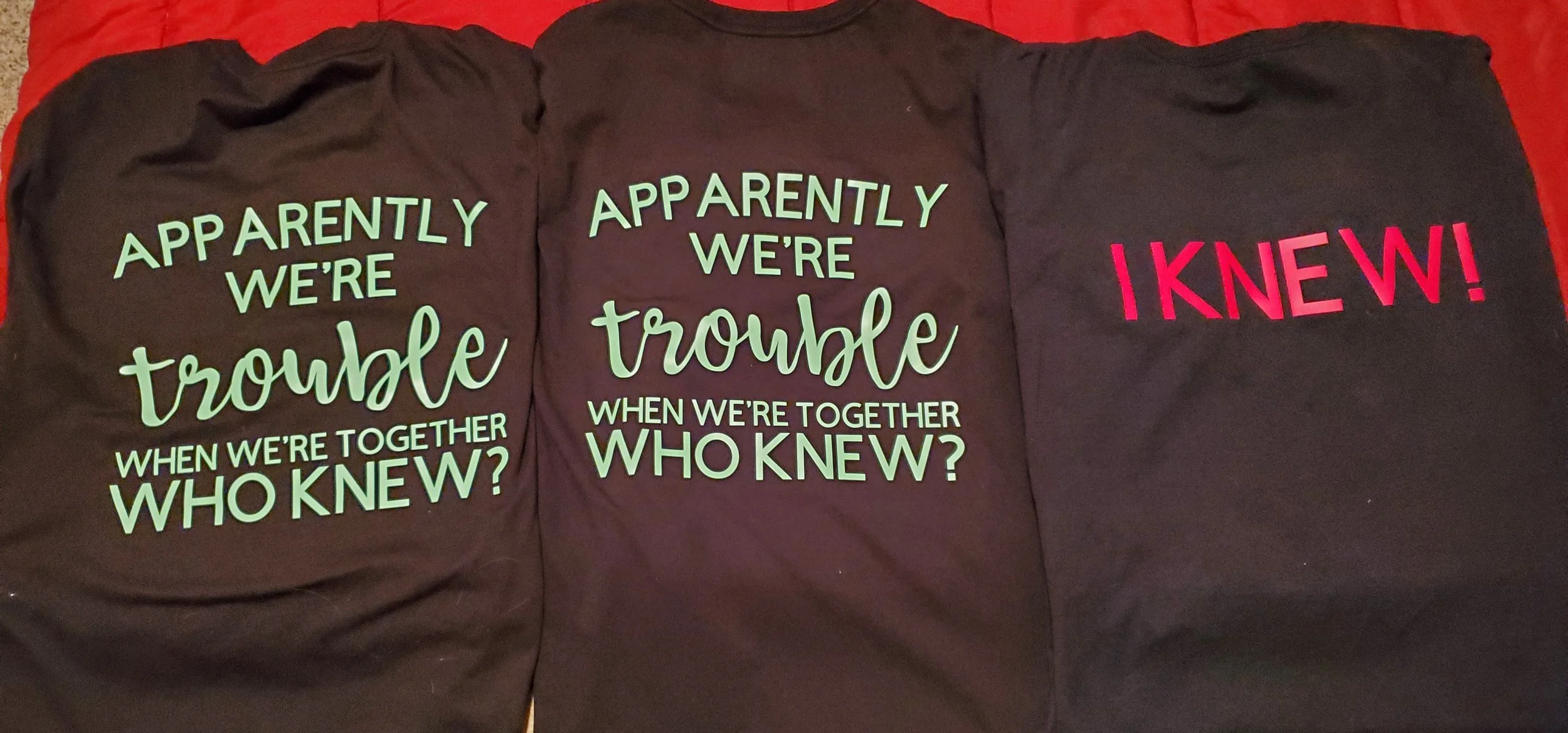 Three black T-shirts with printed phrases, the first two reading "Apparently we're trouble when we're together who knew?" in green and white text, and the third with "I knew!" in red text.