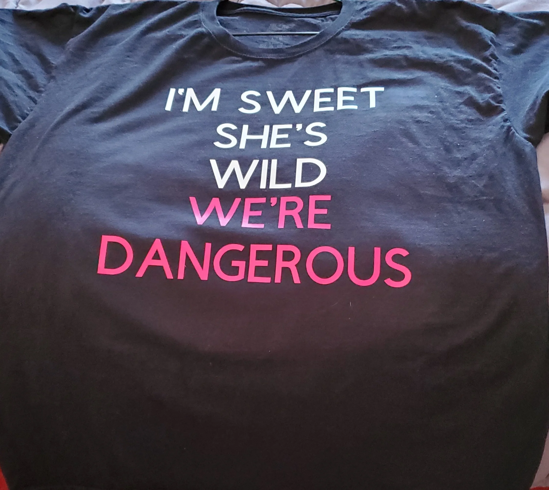 A black T-shirt with text that reads 'I'M SWEET SHE'S WILD WE'RE DANGEROUS'. The text is in white, pink, and purple colors.