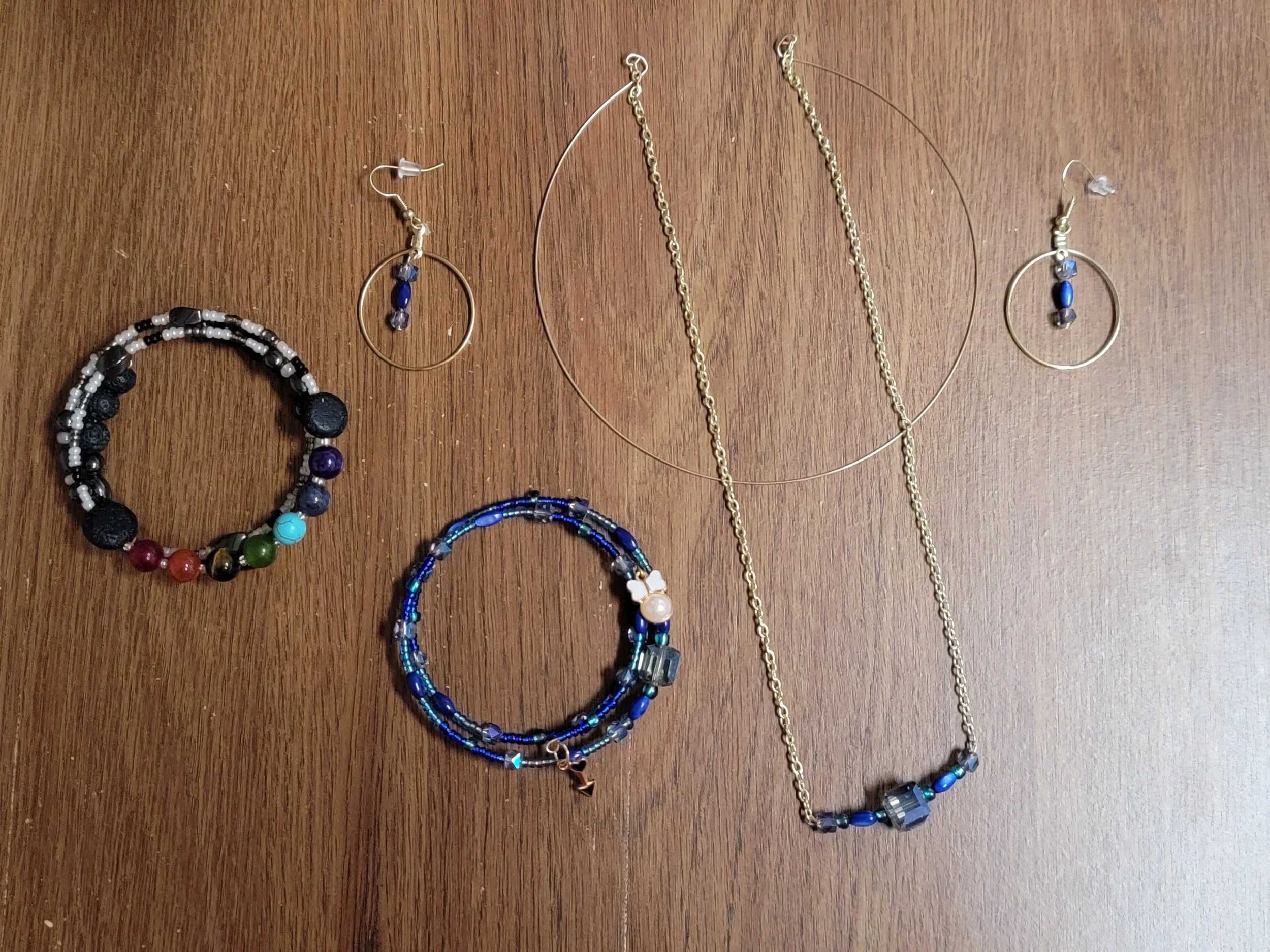 Collection of jewelry including a beaded bracelet, a small hoop earring with blue beads, a large gold hoop earring, a gold necklace, a multi-layered blue beaded bracelet, and a gold chain necklace with blue and gray beads, arranged on a wooden surfac