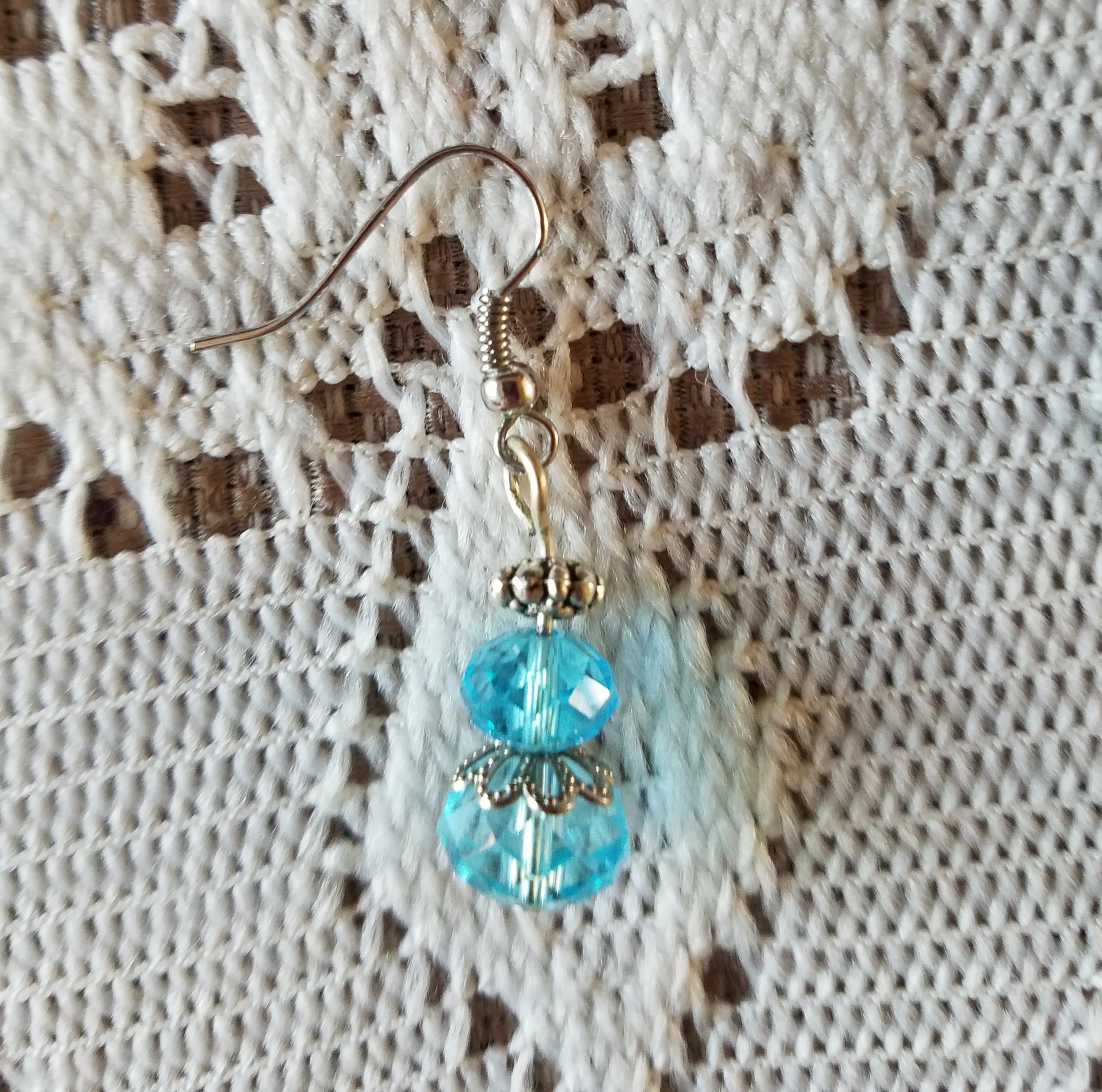 Earring with a silver hook, small silver beads, and a dangling charm featuring two blue faceted beads separated by a silver decorative element, attached to a white crochet fabric background.