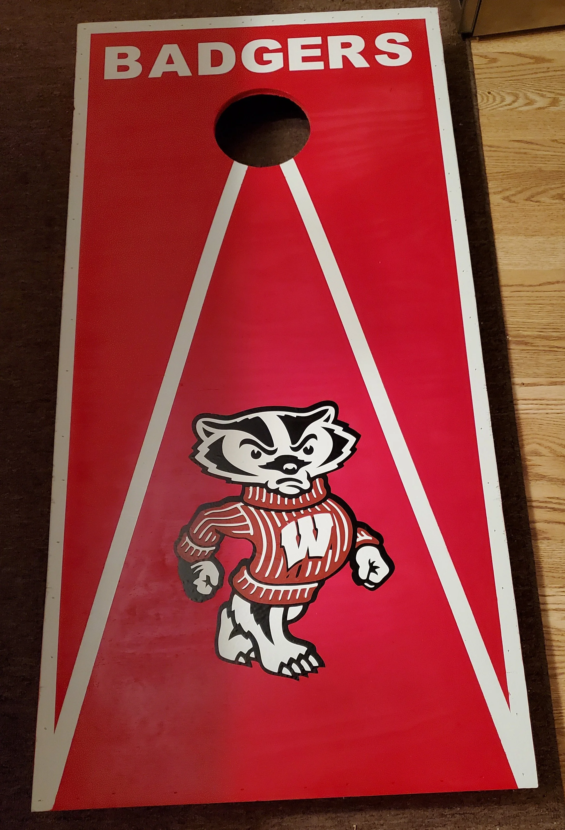 A plastic cornhole game board labeled 'Badgers' with a cartoon badger mascot wearing a red striped sweater with a white 'W' on it, standing in a stance with fists clenched.