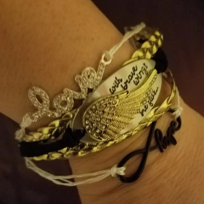 A collection of bracelets, including a black, yellow, and cream braided bracelet, a silver bracelet with the word 'Love' in black, and a gold bracelet with a bejeweled wing design and the phrase 'fight for love' written on it.