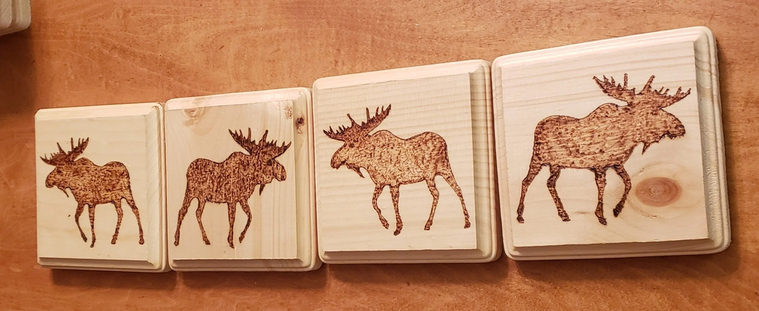 Four wooden tiles with moose silhouettes burned into the wood surface, arranged on a wooden table.
