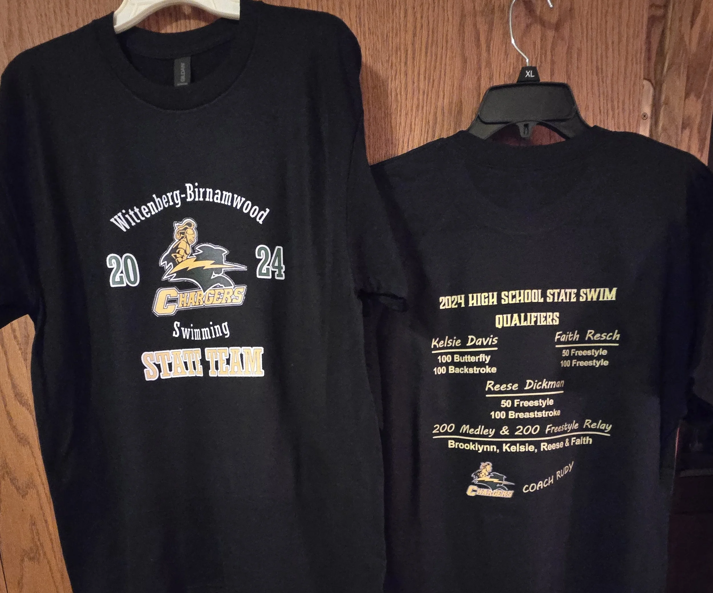 Black T-shirts hanging on a wooden wall, one with a Cleveland Chargers logo and the other with text about high school swim qualifiers.