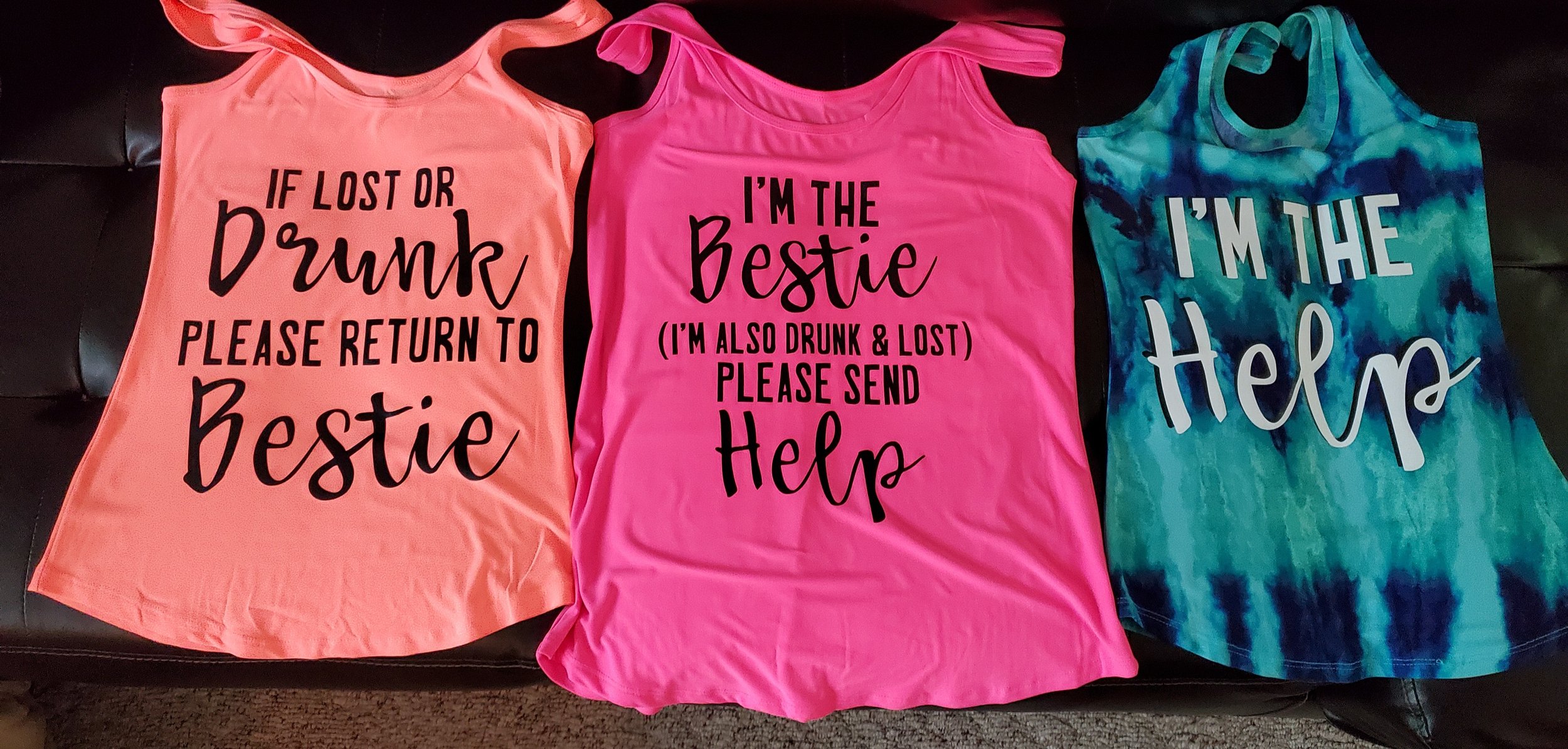 Three tank tops laying flat. The first is peach with black text saying, "If lost or drunk please return to Bestie." The second is pink with black text saying, "I'm the Bestie (I'm also drunk & lost) Please send Help." The third is blue and green tie-