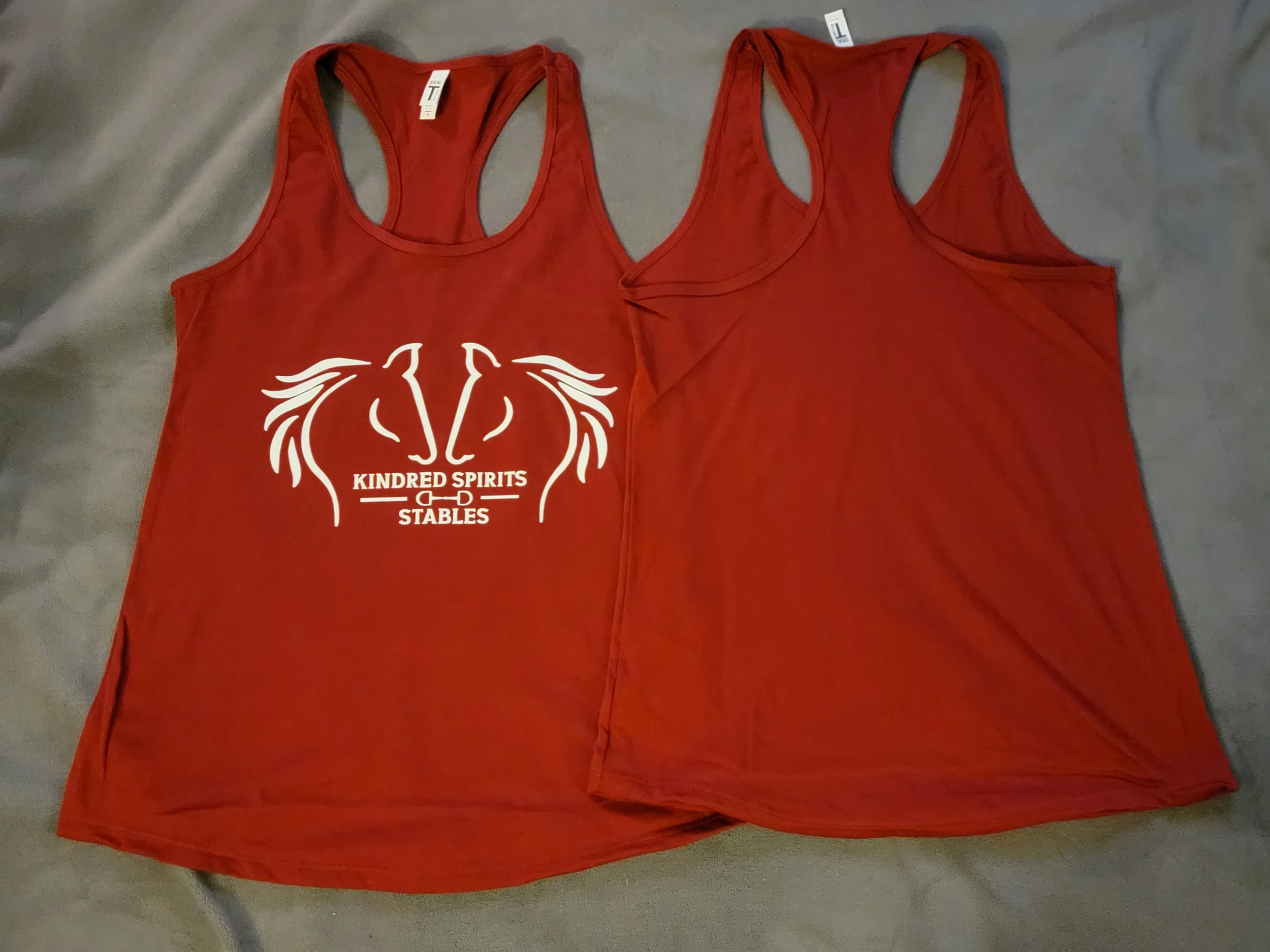 Two red tank tops with racerback straps lying flat on a grey surface, one with white racing logo and text reading 'Kindred Spirits Stables'.