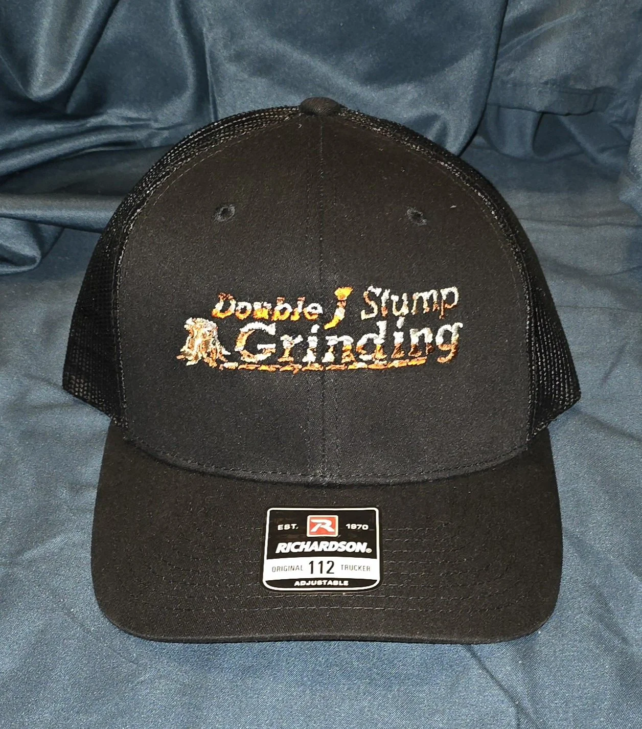 Black trucker baseball cap with embroidered text that reads 'Double J Stump Grinding' and a small image of a tree on the left side of the text. The cap has a mesh back and a sticker on the brim.