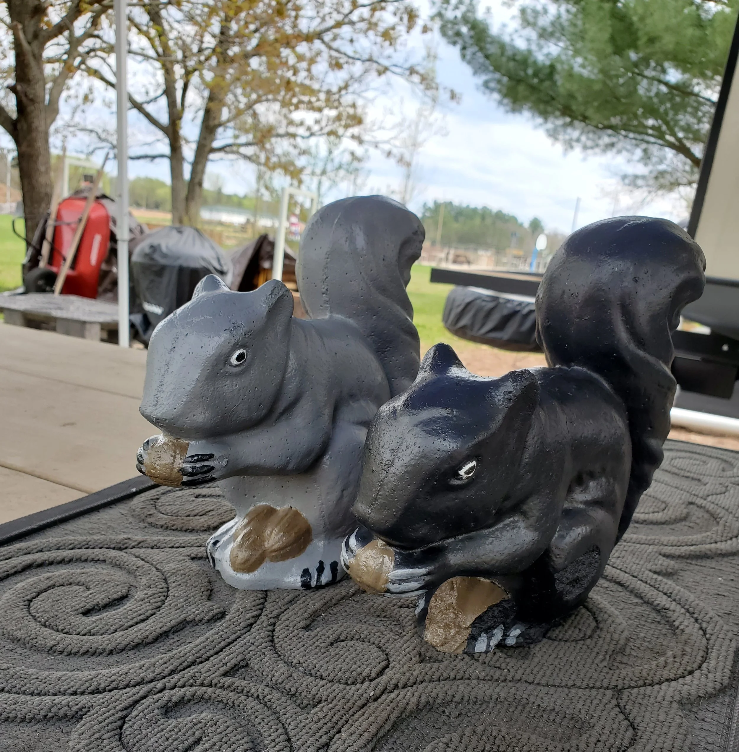 Two squirrel figurines, one gray and one black, sitting on a doormat with a textured swirl pattern, outdoors on a wooden table.