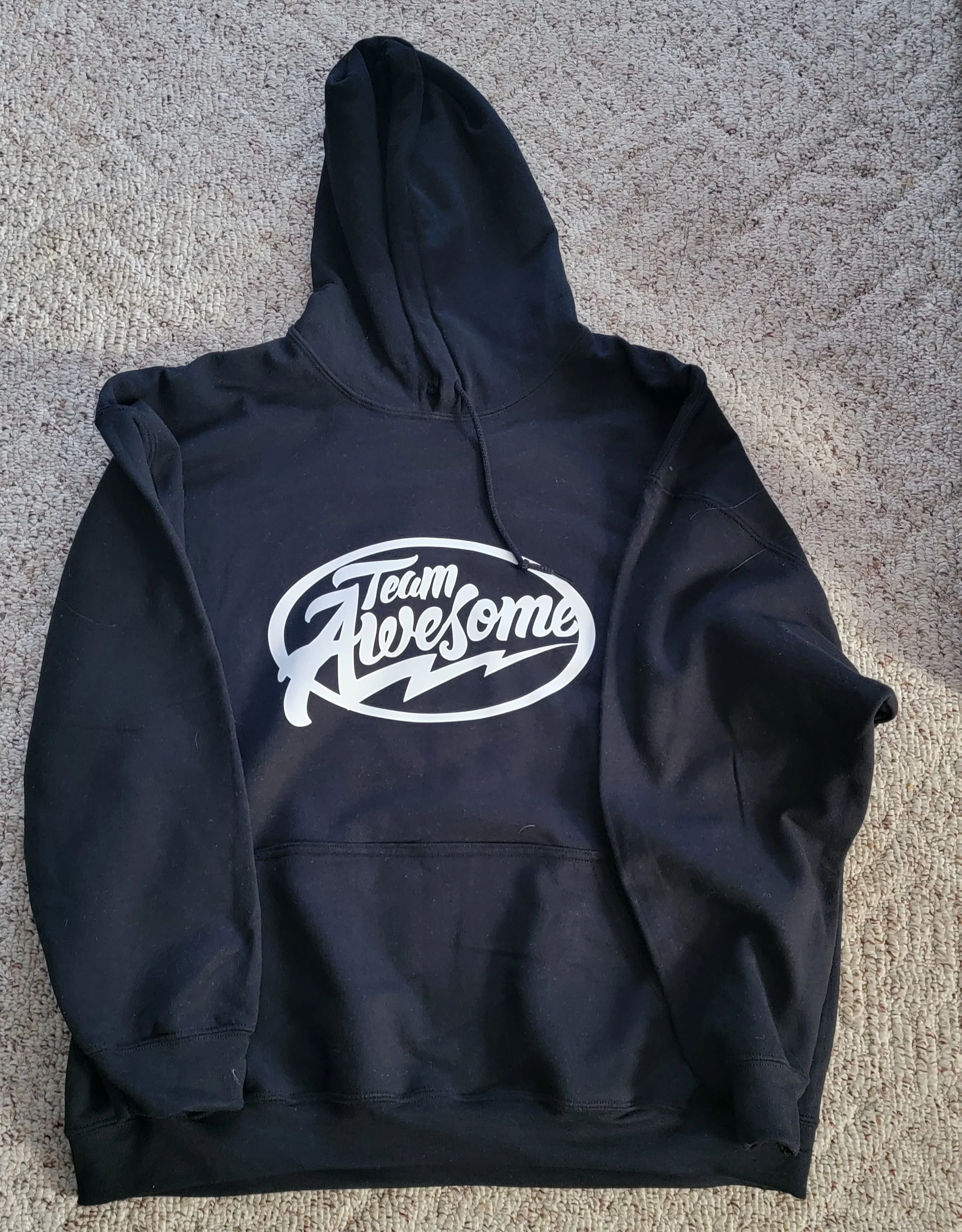 Black hoodie with a white oval logo that says 'Team Awesome' on the front, laid flat on a beige carpet.