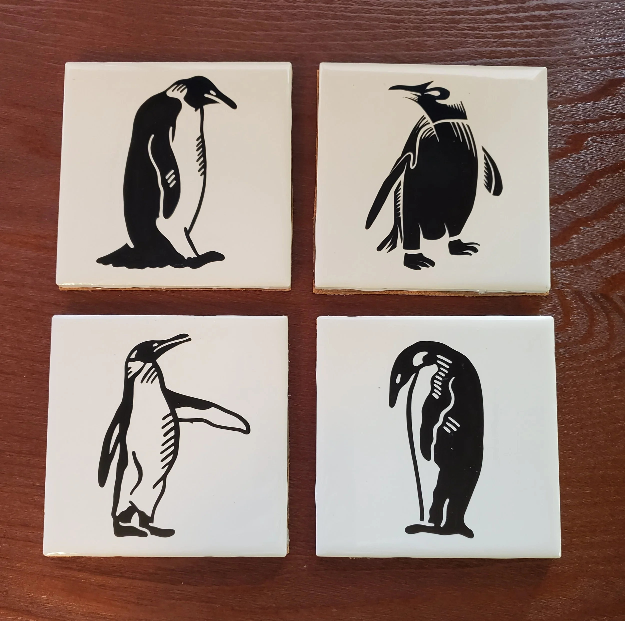 Four black and white illustrated tiles featuring different penguin sketches, arranged in a 2x2 grid on a wooden surface.